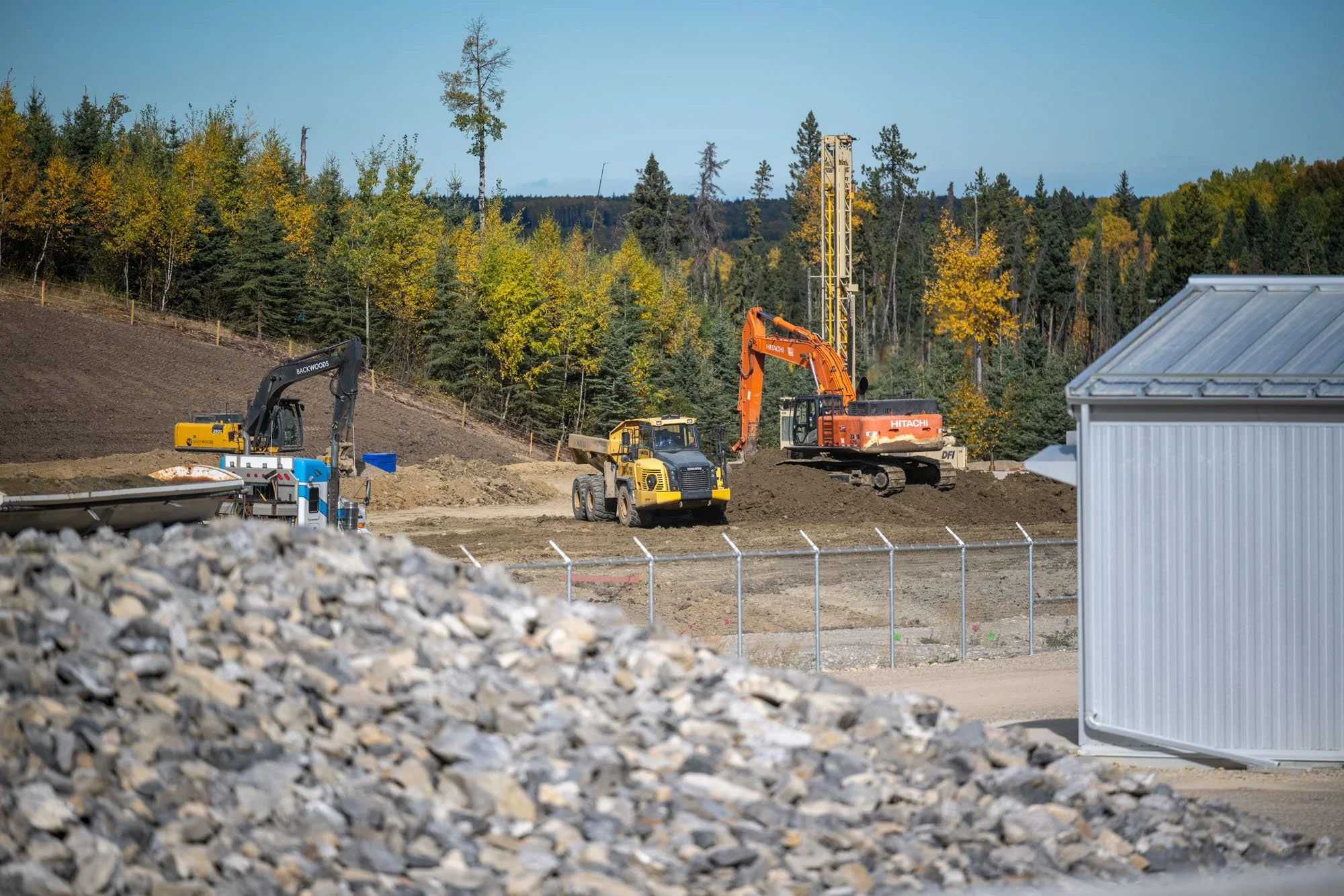 Construction site with excavators and bulldozer working on land, surrounded by forested area, with a metal shed on the right and a pile of rocks in the foreground.