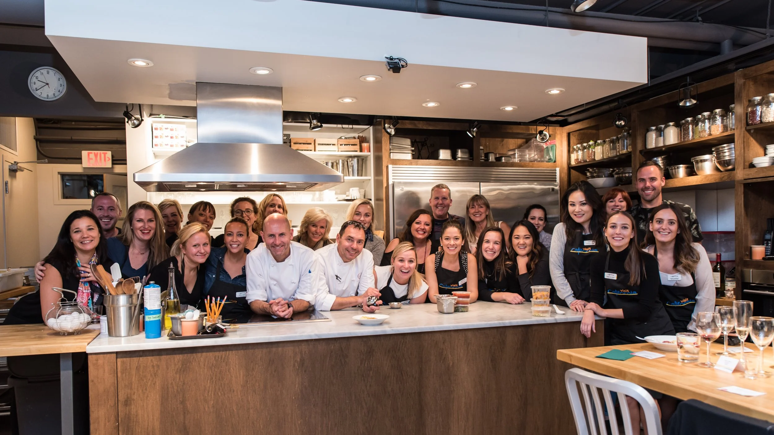 Group of people gathered in a kitchen, smiling for a photo, with some wearing aprons and chef's coats.