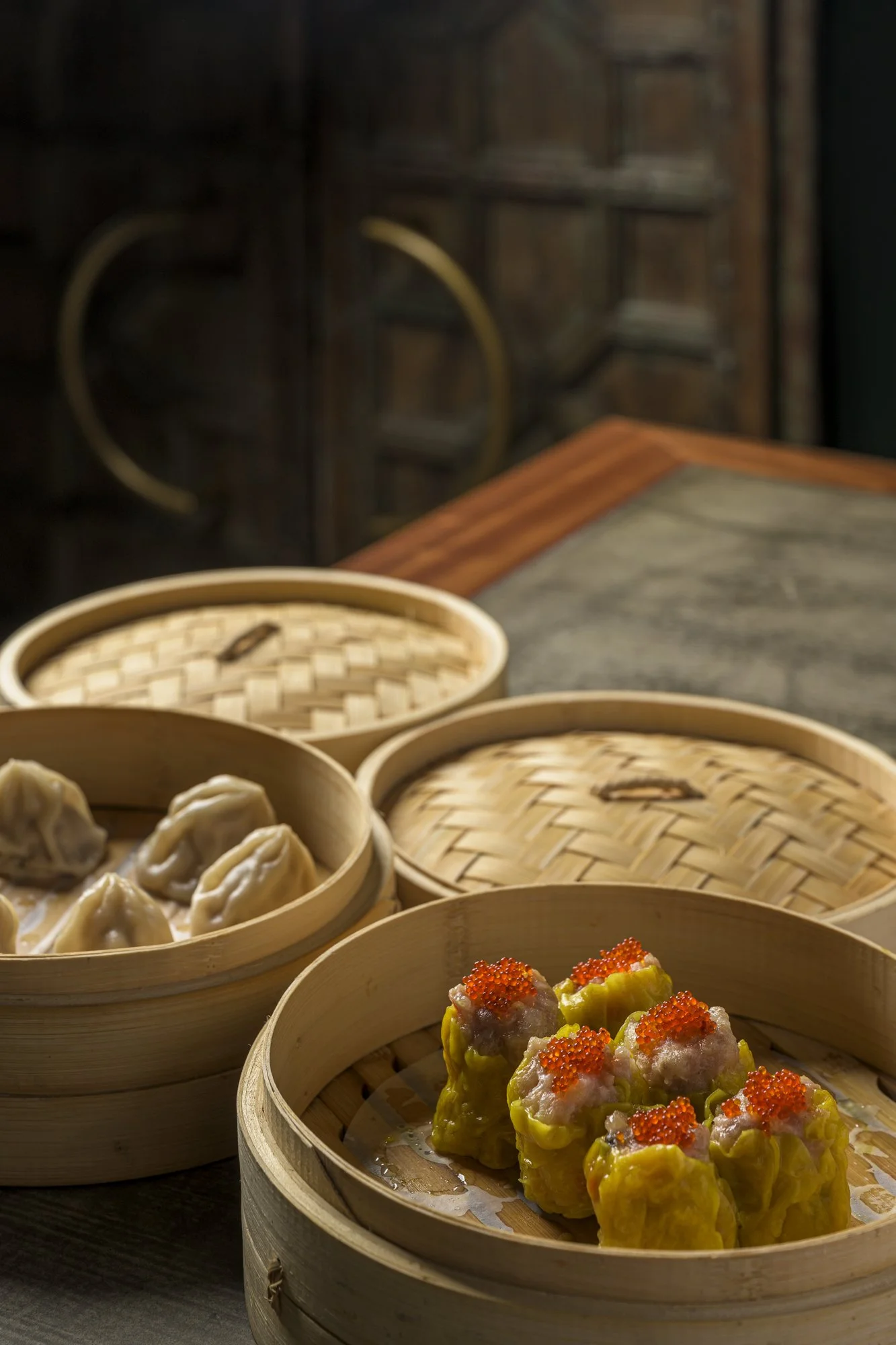 A close-up of a bamboo steamer basket filled with yellow dumplings topped with orange fish roe, sitting on a table with other stacked bamboo steamers containing additional dumplings.