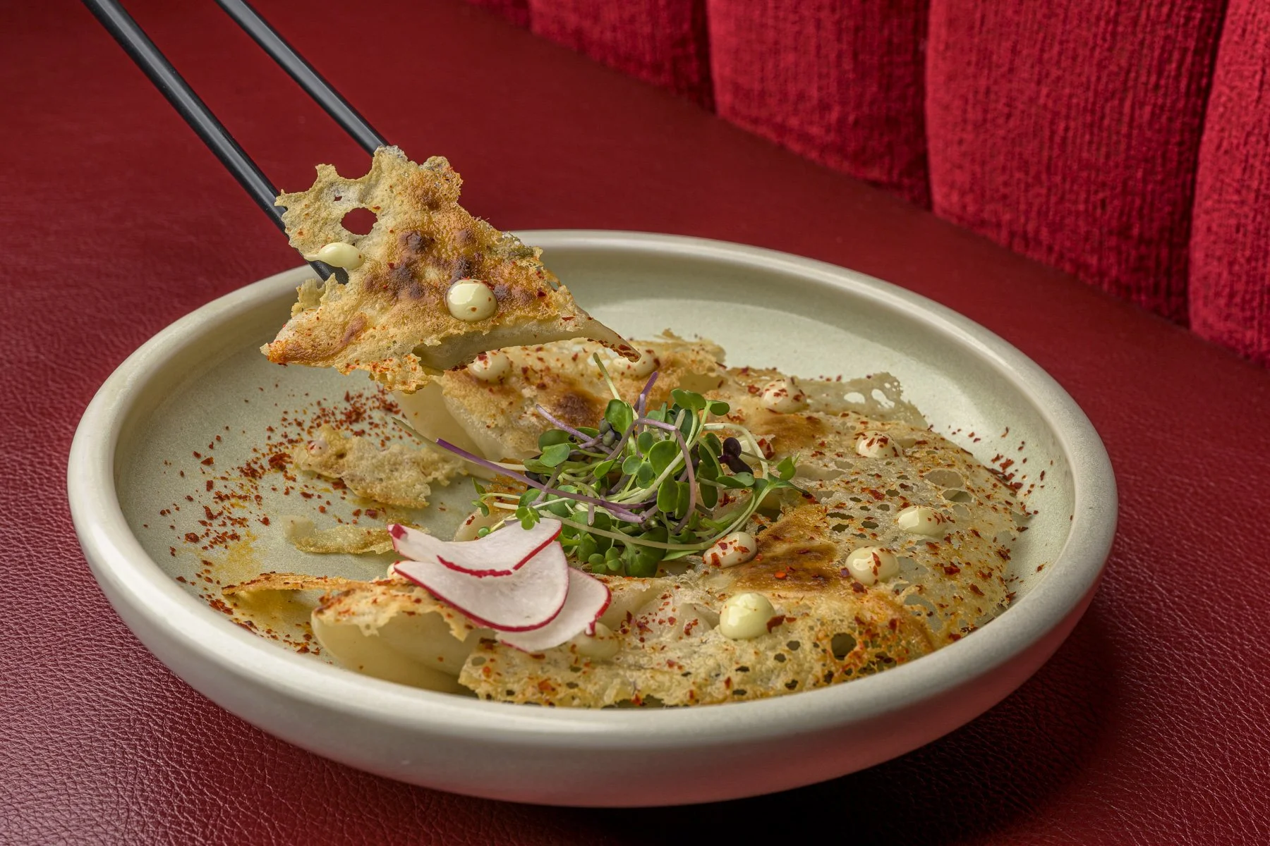 A plate of Japanese gyoza dumplings garnished with microgreens, thinly sliced radishes, and drizzled with white sauce, on a red textured surface.