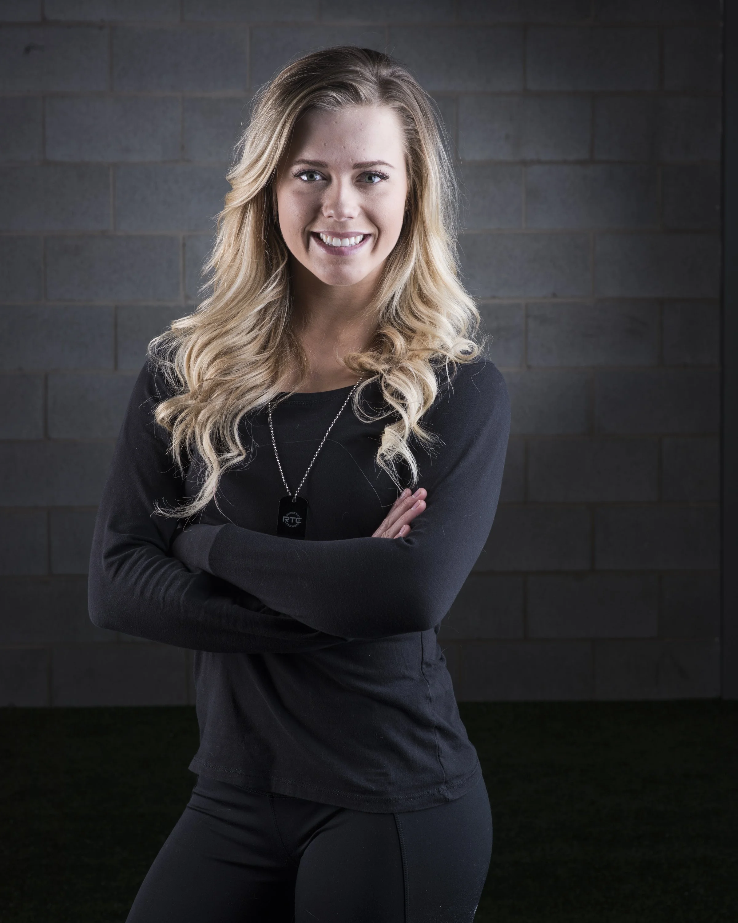 Young woman with blonde hair smiling and crossing her arms, wearing black clothing and a military dog tag necklace, standing against a dark brick wall.
