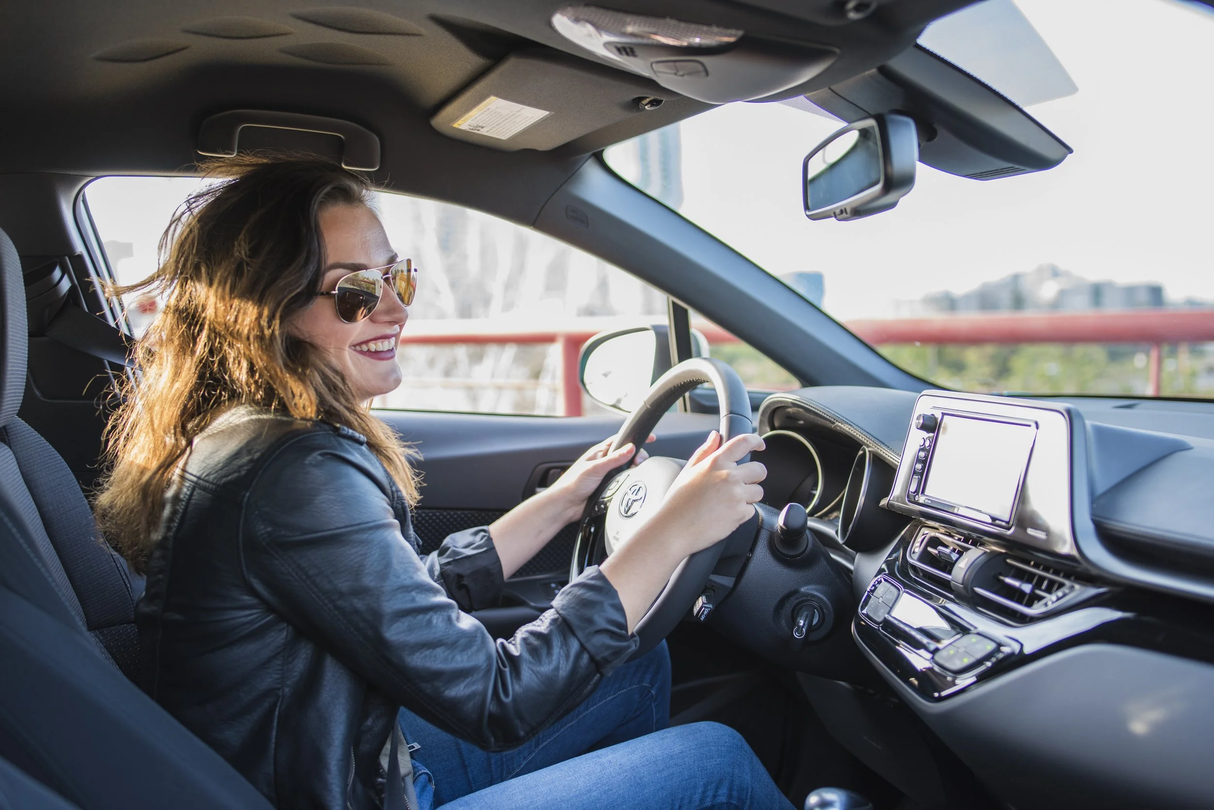 Woman driving a car, wearing sunglasses, black leather jacket, and smiling, with cityscape visible through the windshield.