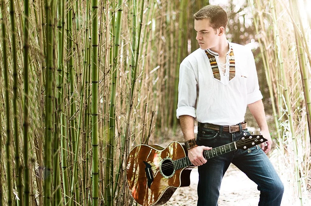 Young man with short dark hair, wearing a white shirt and jeans, standing in a bamboo forest holding an acoustic guitar.