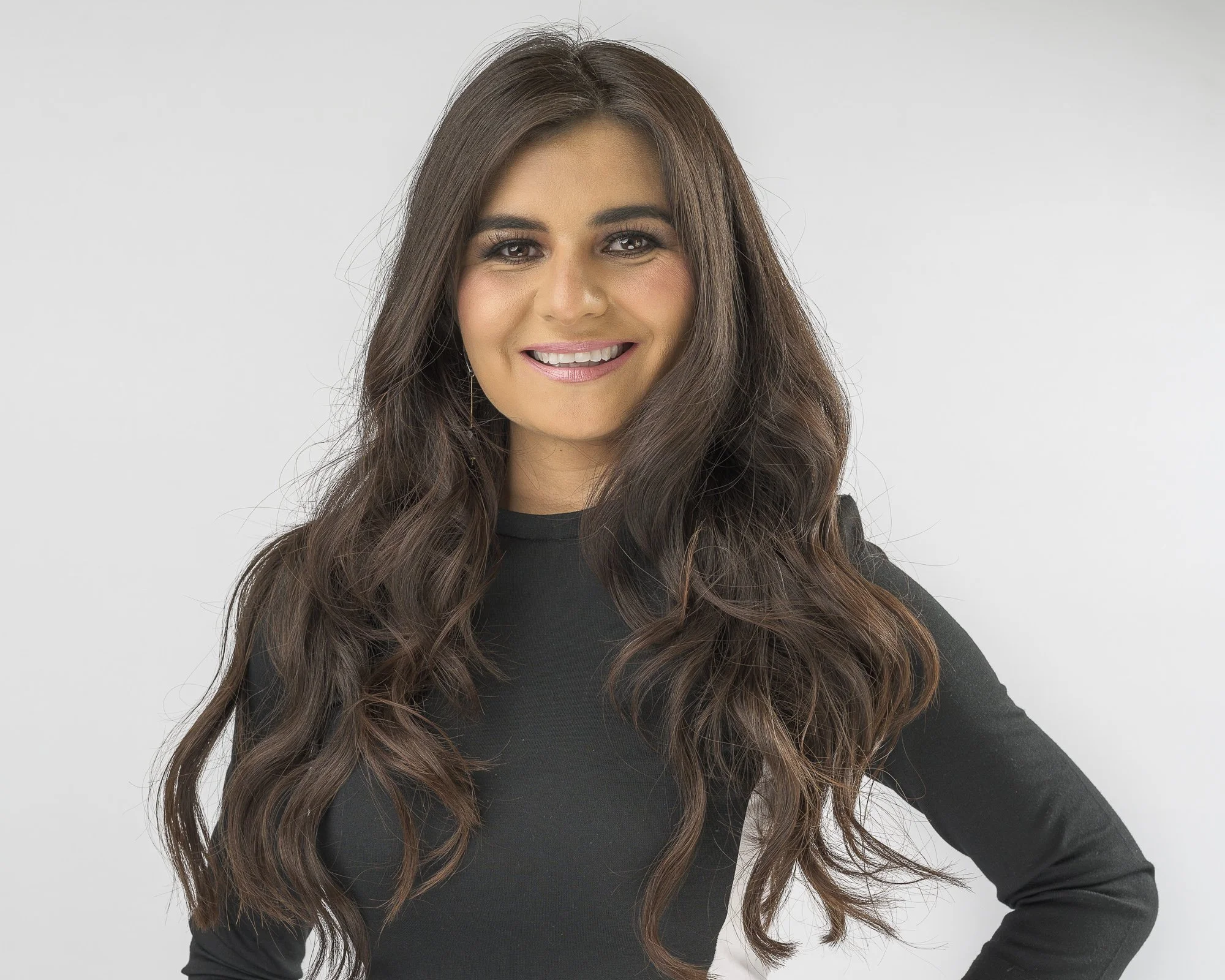 A woman with long, wavy brown hair wearing a black top, smiling, with a plain light gray background.