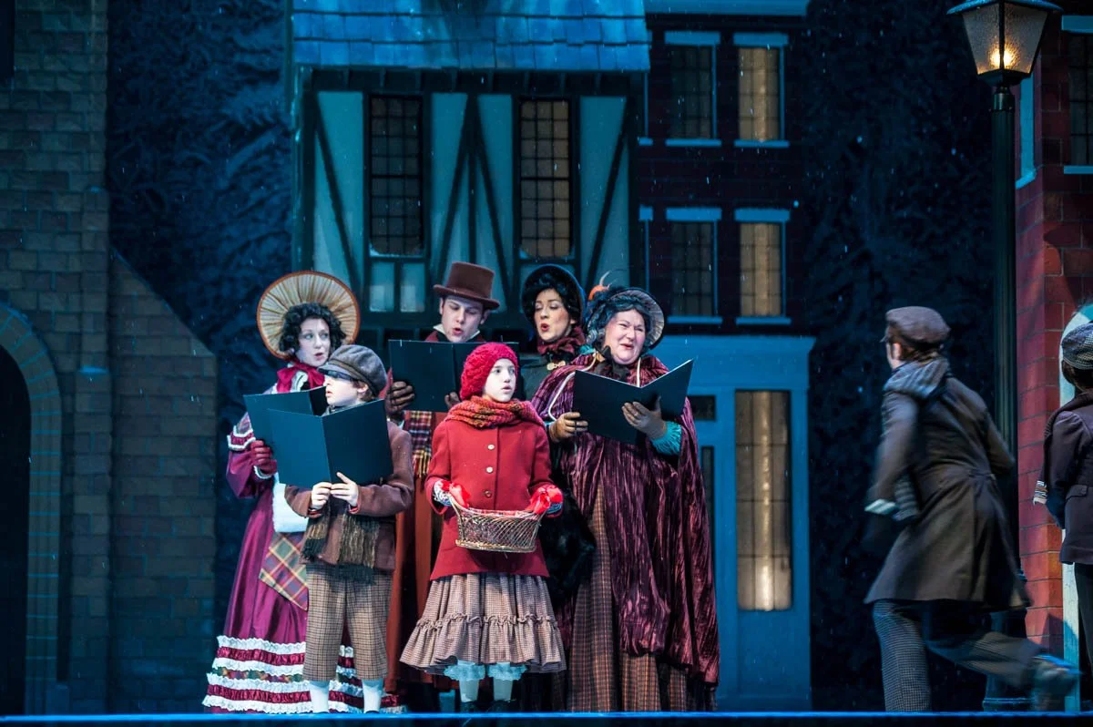 People dressed in Victorian costumes singing on stage, with some looking at sheet music, outdoors at night, illuminated by a streetlamp.