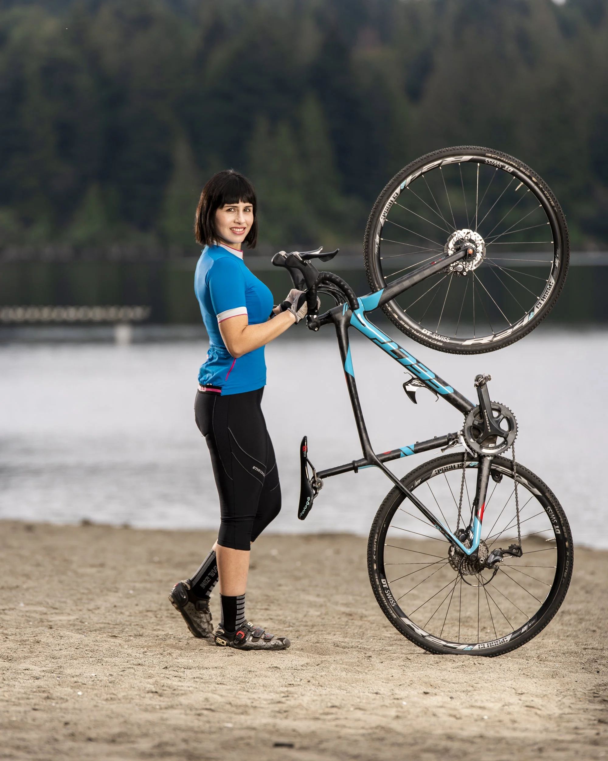 A woman in cycling gear holding a mountain bike with one wheel raised, standing on a sandy beach near water with trees in the background.