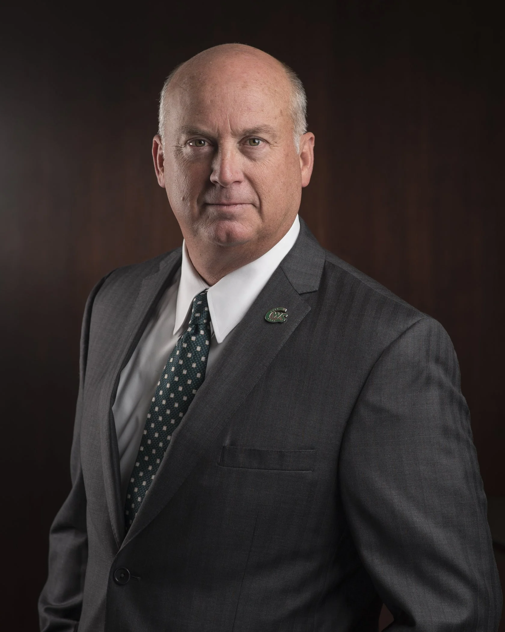 A middle-aged man with a bald head in a dark suit, white shirt, and polka-dot tie, posing against a dark background