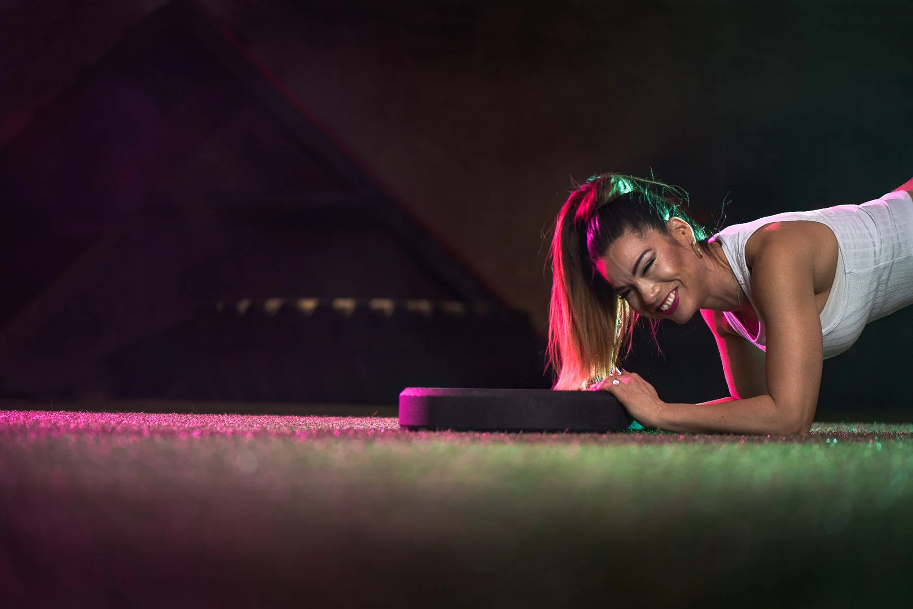 A woman is exercising on a fitness mat, smiling with her eyes closed, under colorful stage lighting.