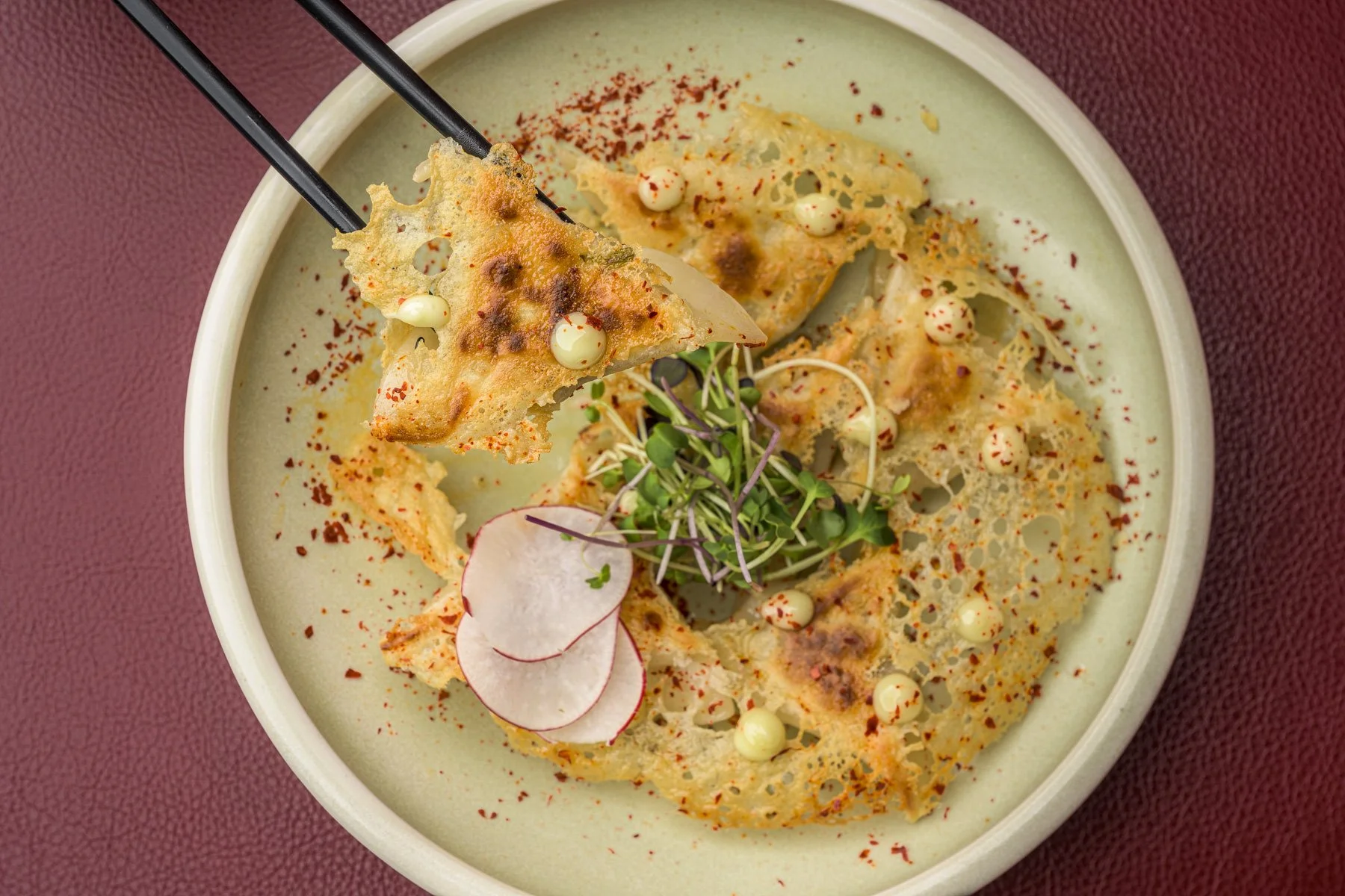 A bowl of Asian-inspired soup topped with crispy wonton chips, radish slices, microgreens, and white sauce pearls, with a spoon lifting a wonton chip.