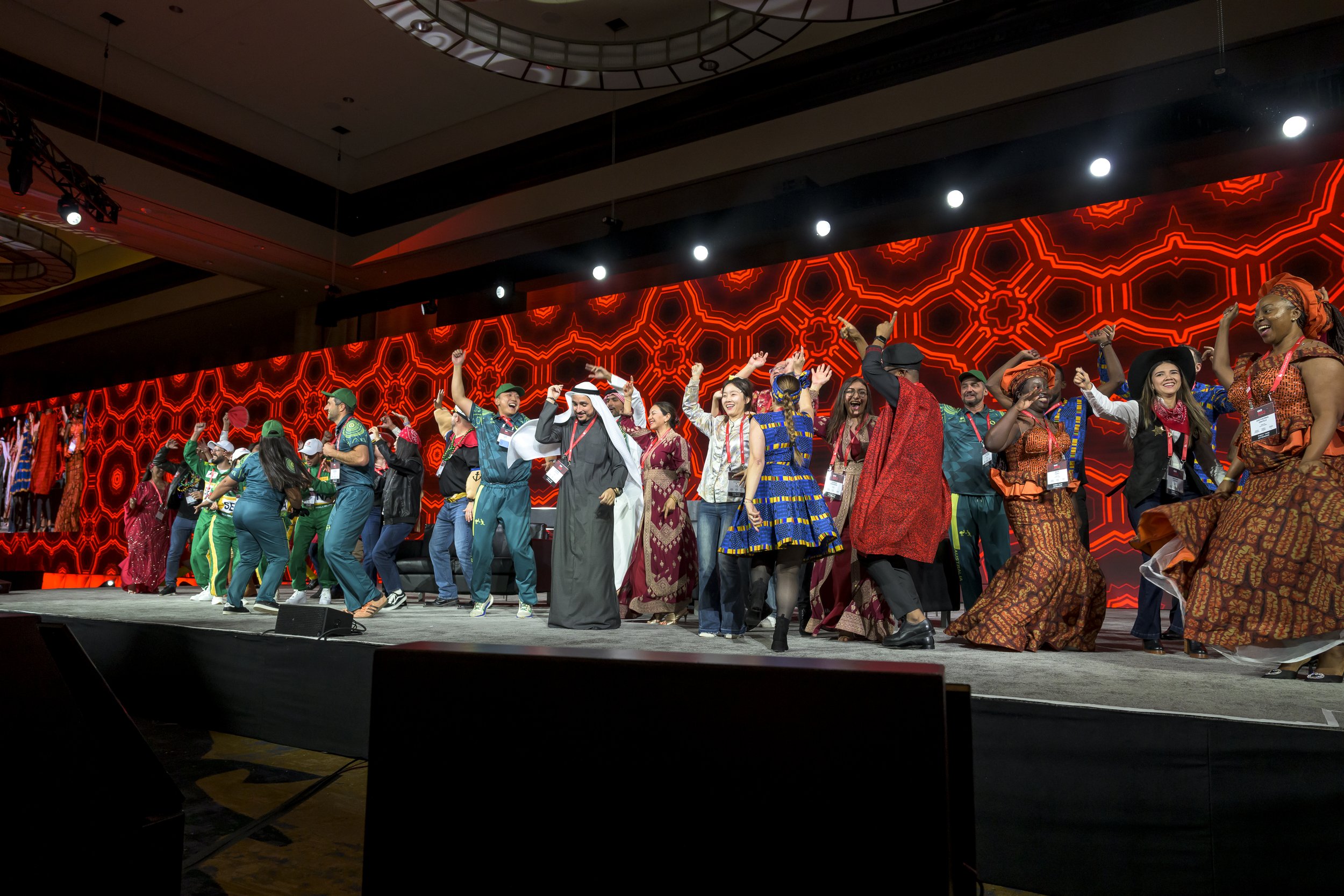 A diverse group of people dancing and celebrating on a stage with a bright red geometric patterned backdrop at a conference or event.