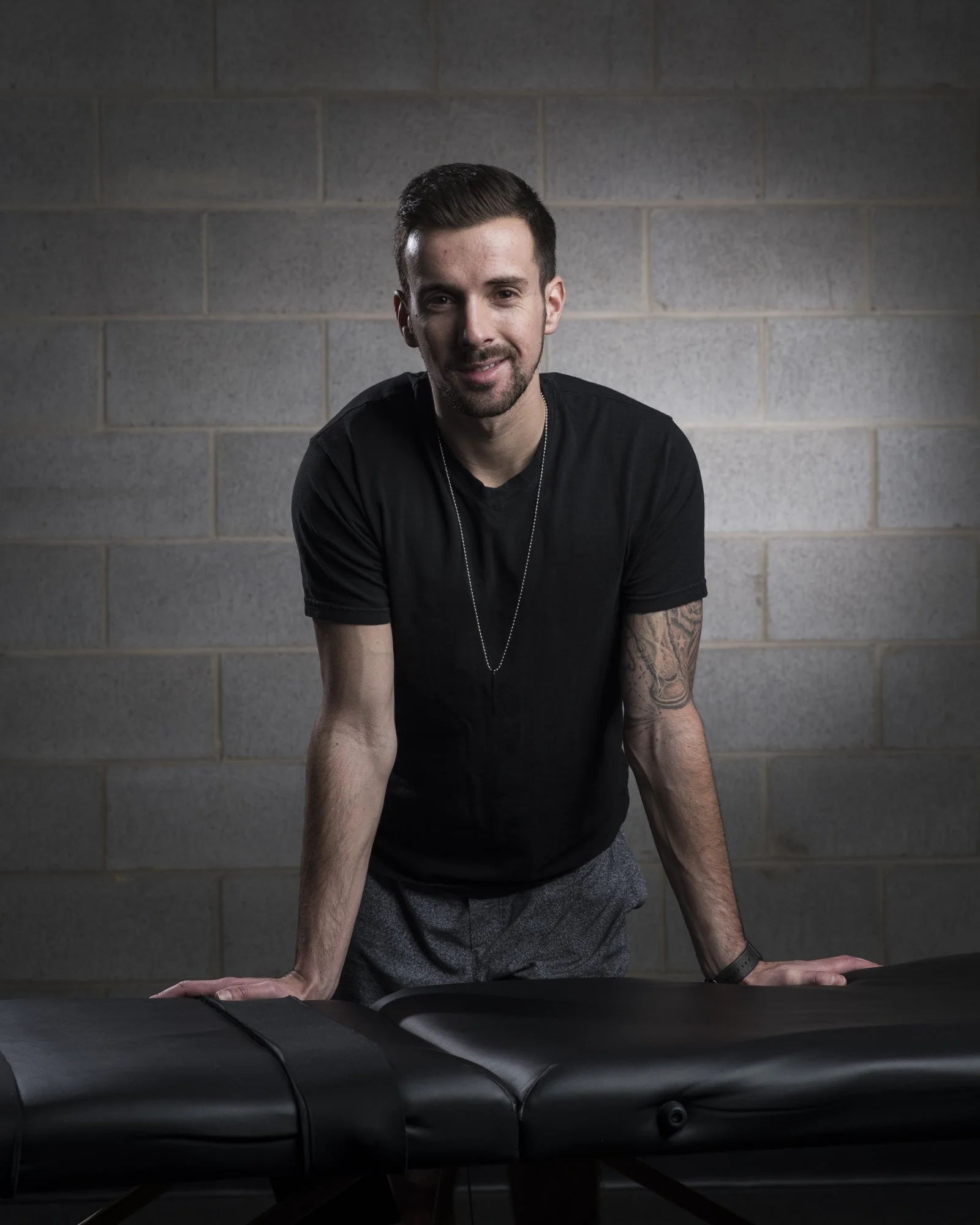 A man with dark hair, beard, and tattoos on his left arm, wearing a black t-shirt and a necklace, leaning on a massage table against a gray brick wall.