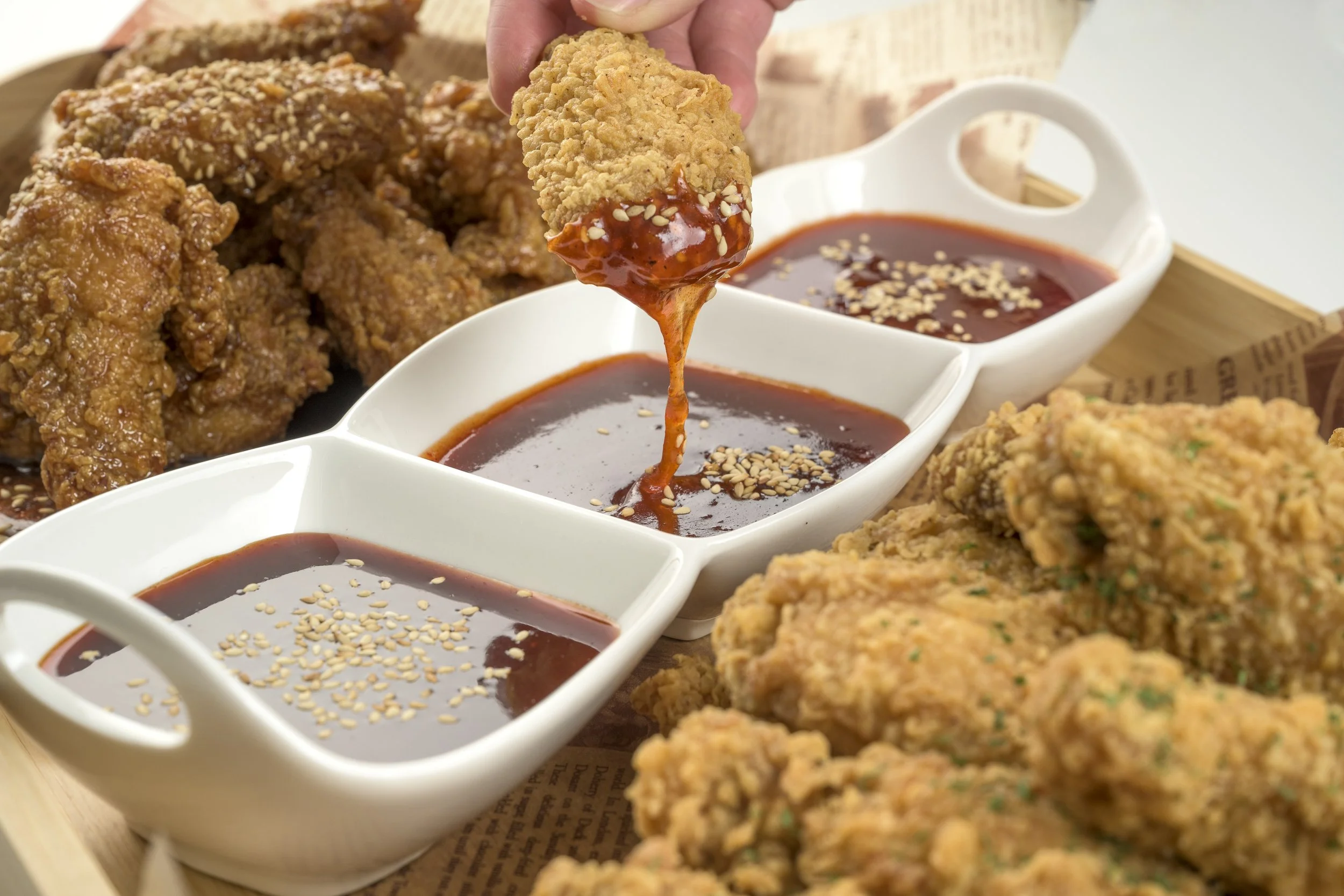 Fried chicken pieces with three containers of dipping sauces, one of which is being spooned with sauce. The sauces are garnished with sesame seeds, and there is a serving of fried chicken in the foreground and background.