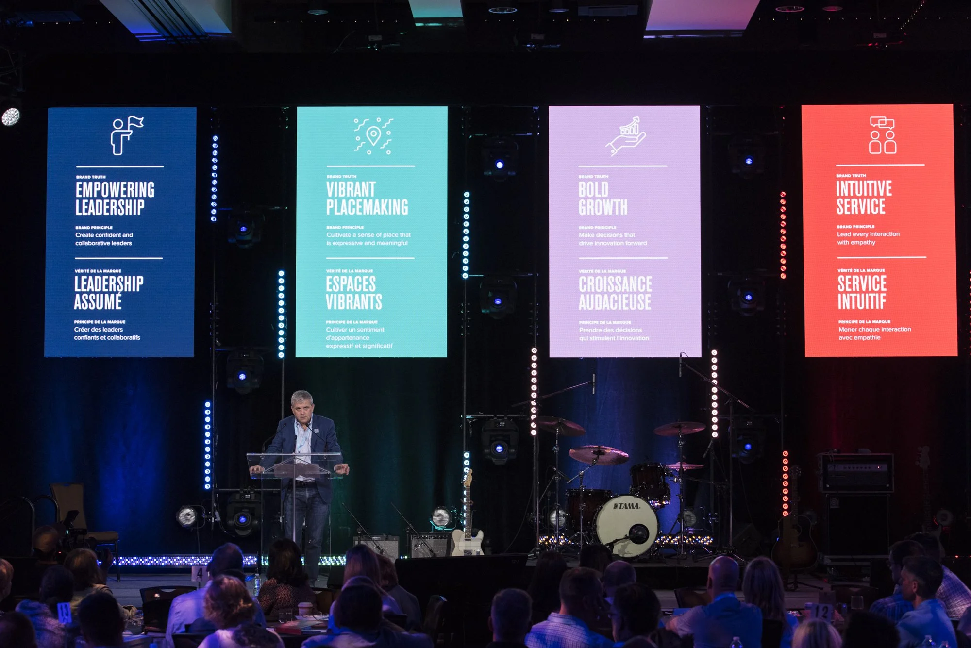 A man presenting on stage at a conference with large digital screens behind him displaying leadership principles in English and French. The screens show blue, turquoise, purple, and red backgrounds with icons and text about empowering leadership, vib