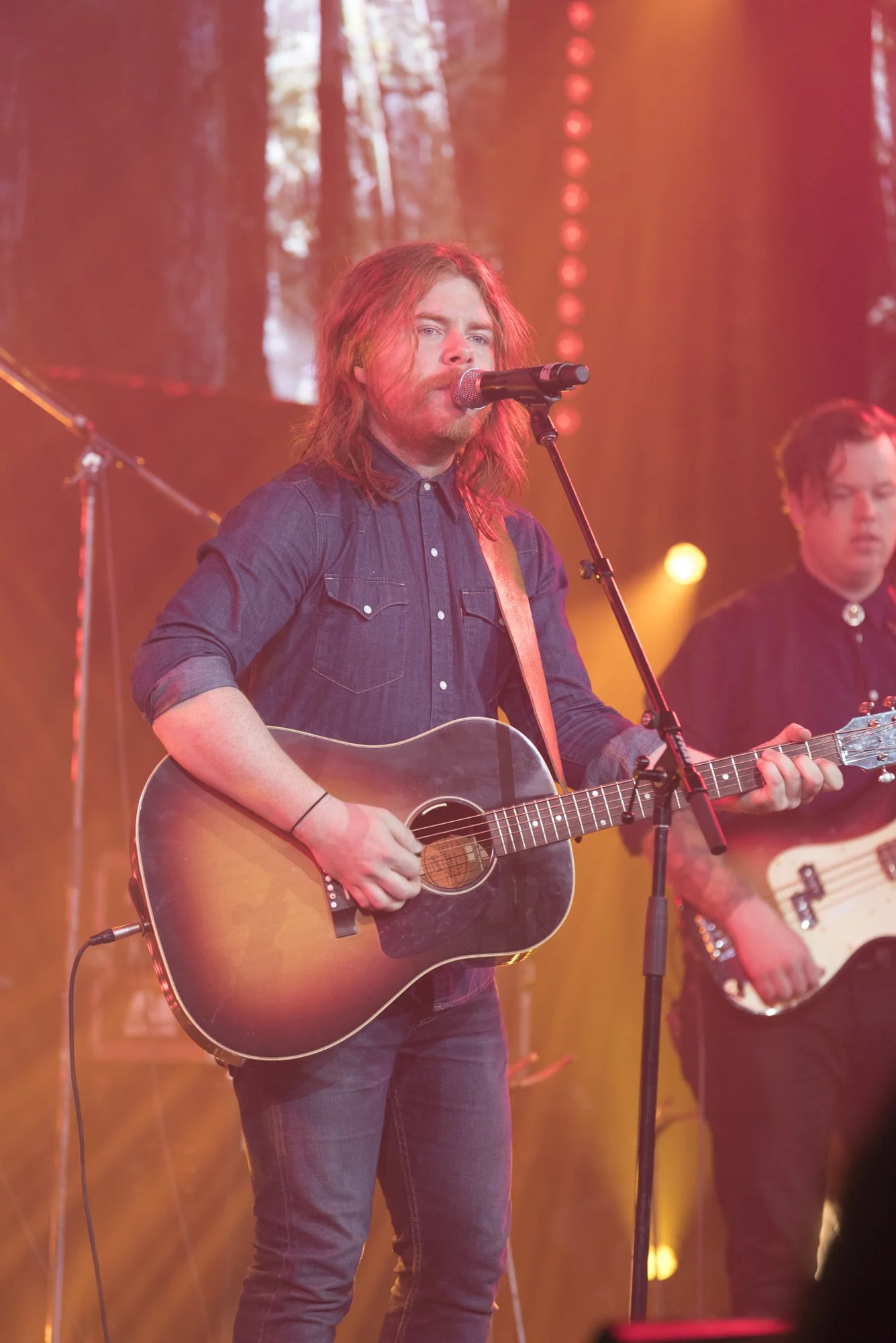 A male musician with long red hair singing into a microphone and playing an acoustic guitar on stage with colorful lights.