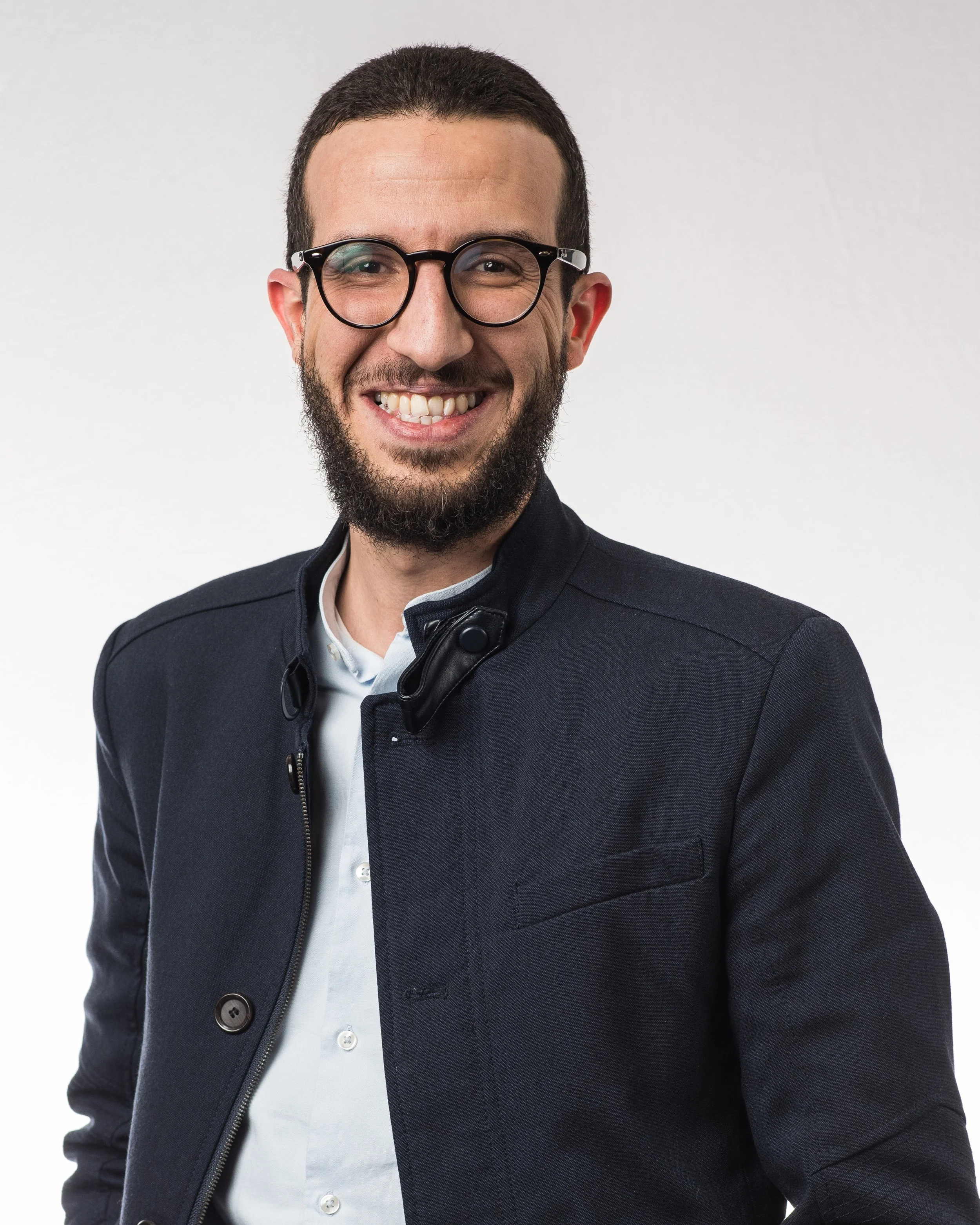 A smiling man with glasses, dark hair, and a beard, wearing a dark jacket and a white shirt, standing against a plain background.
