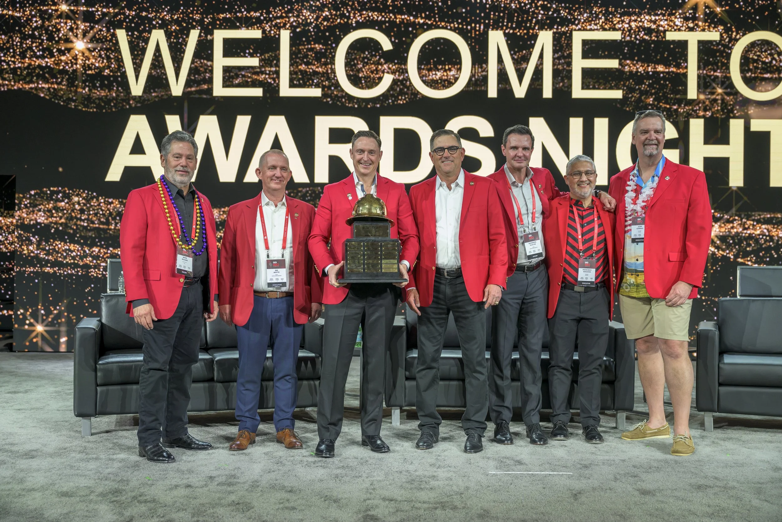 A group of seven men on stage at an awards night, posing with a trophy. They are all wearing red blazers and smiling. The background has a large display with the text 'WELCOME TO AWARDS NIGHT' and decorative lights.