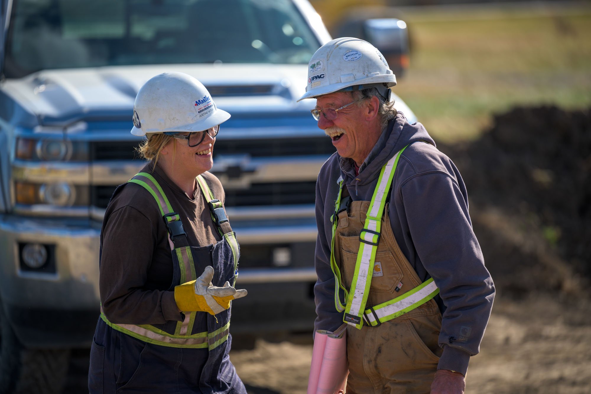 Two construction workers, a woman and a man, are smiling and talking to each other on a work site. They are wearing white safety helmets, safety vests, and work gloves, with a large vehicle in the background.