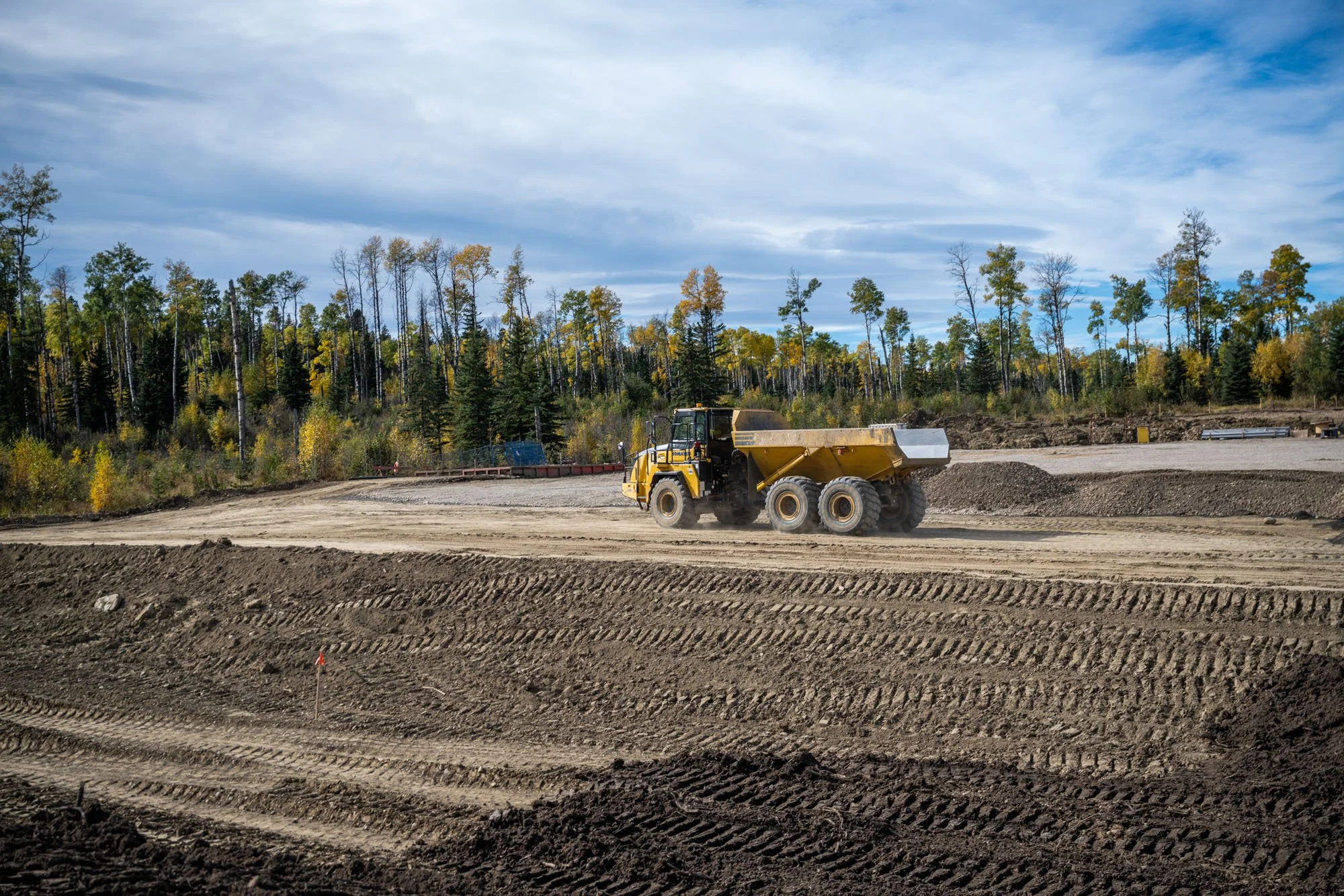 Construction site with a yellow dump truck driving on dirt, surrounded by trees under a partly cloudy sky.