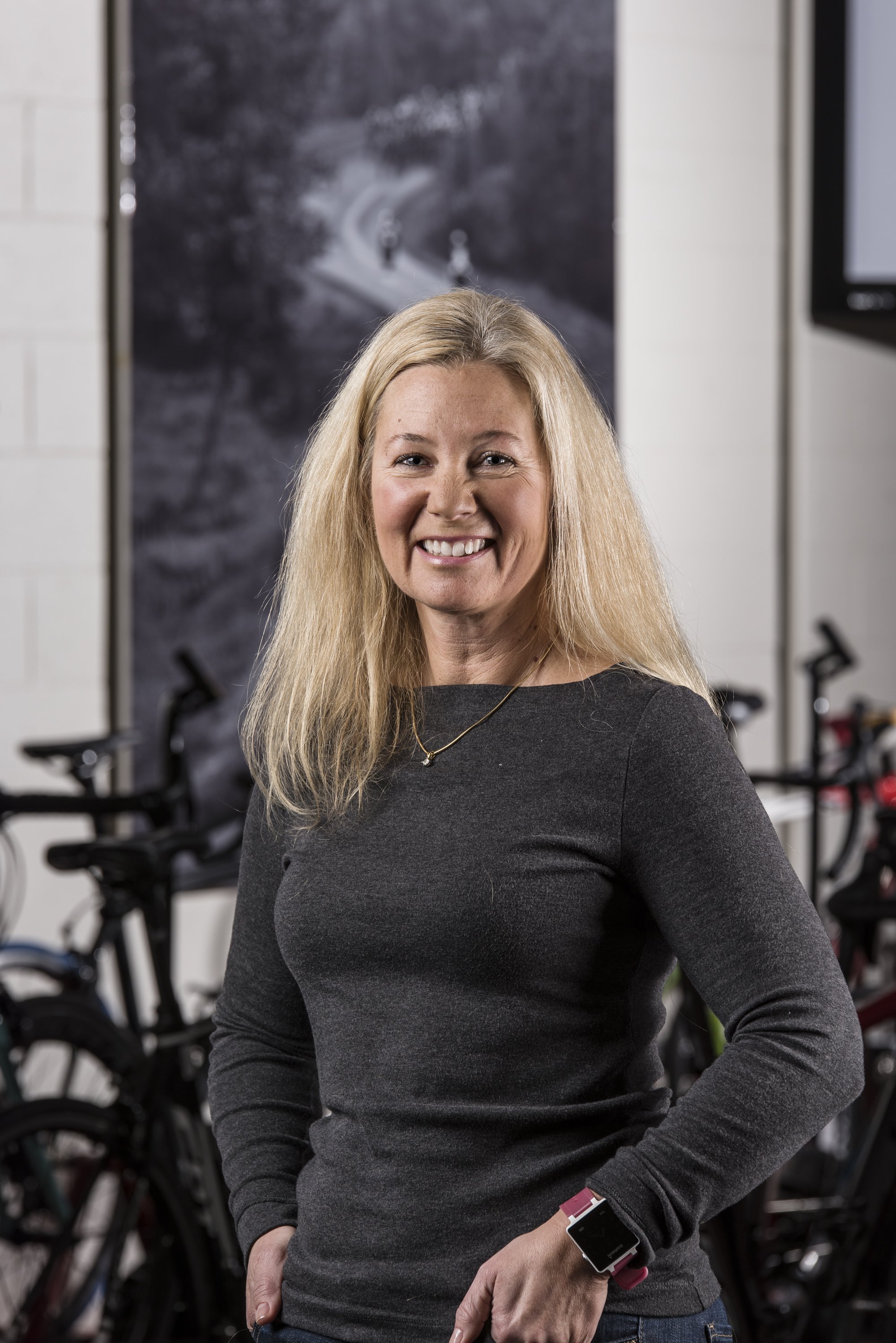 A smiling woman with long blonde hair standing in front of bicycles in a cycling shop or studio.