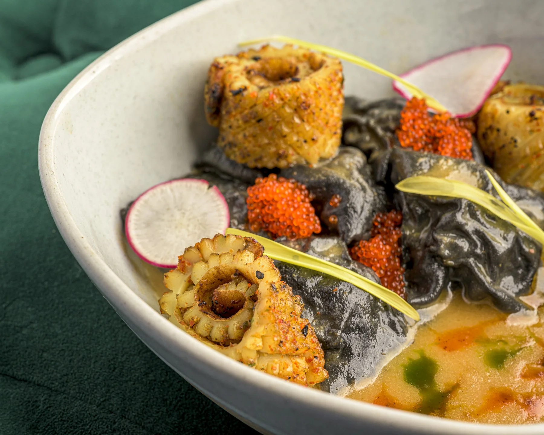 A bowl of gourmet seafood dish with octopus tentacles, black pasta, and garnishes like radish slices, red roe, and octopus pieces, decorated with yellow edible flowers.