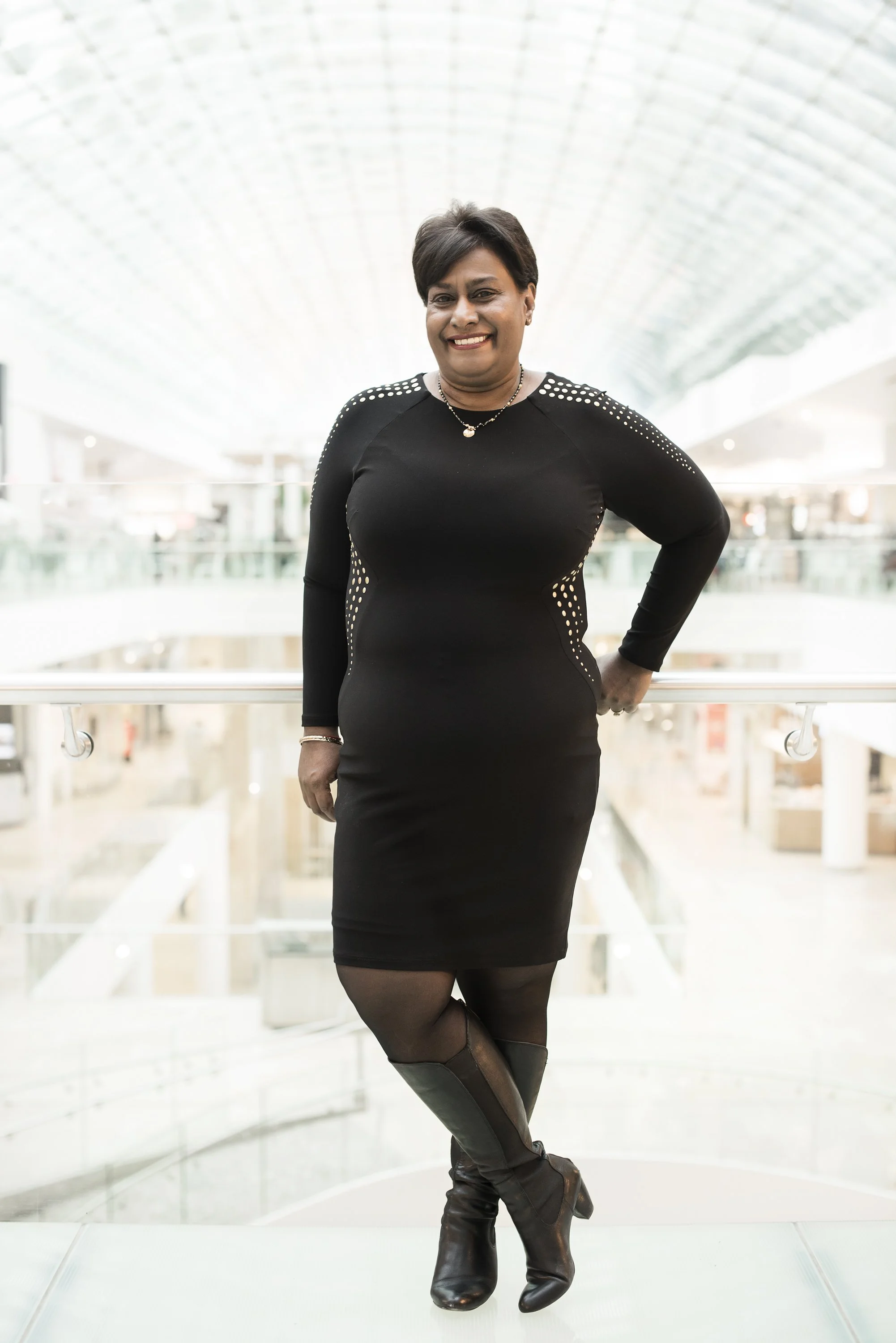 Woman in a black dress standing in a modern indoor space, smiling with one hand on her hip.