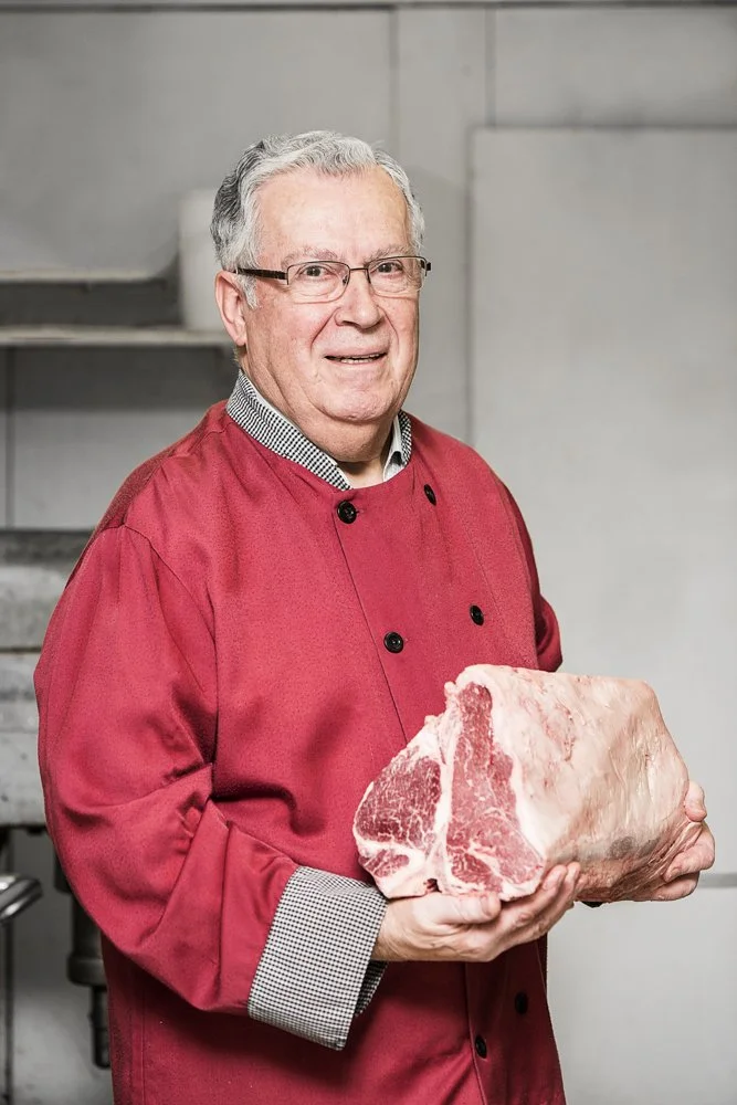 An elderly man in a red chef's coat holding a large piece of raw beef in a kitchen setting.
