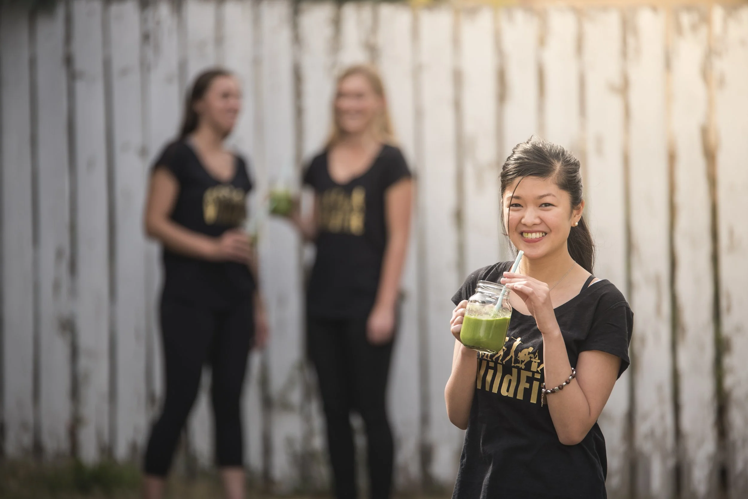 Smiling woman in black T-shirt holding a green smoothie in a mason jar, with two women in black T-shirts in the background, standing in front of a wooden fence.