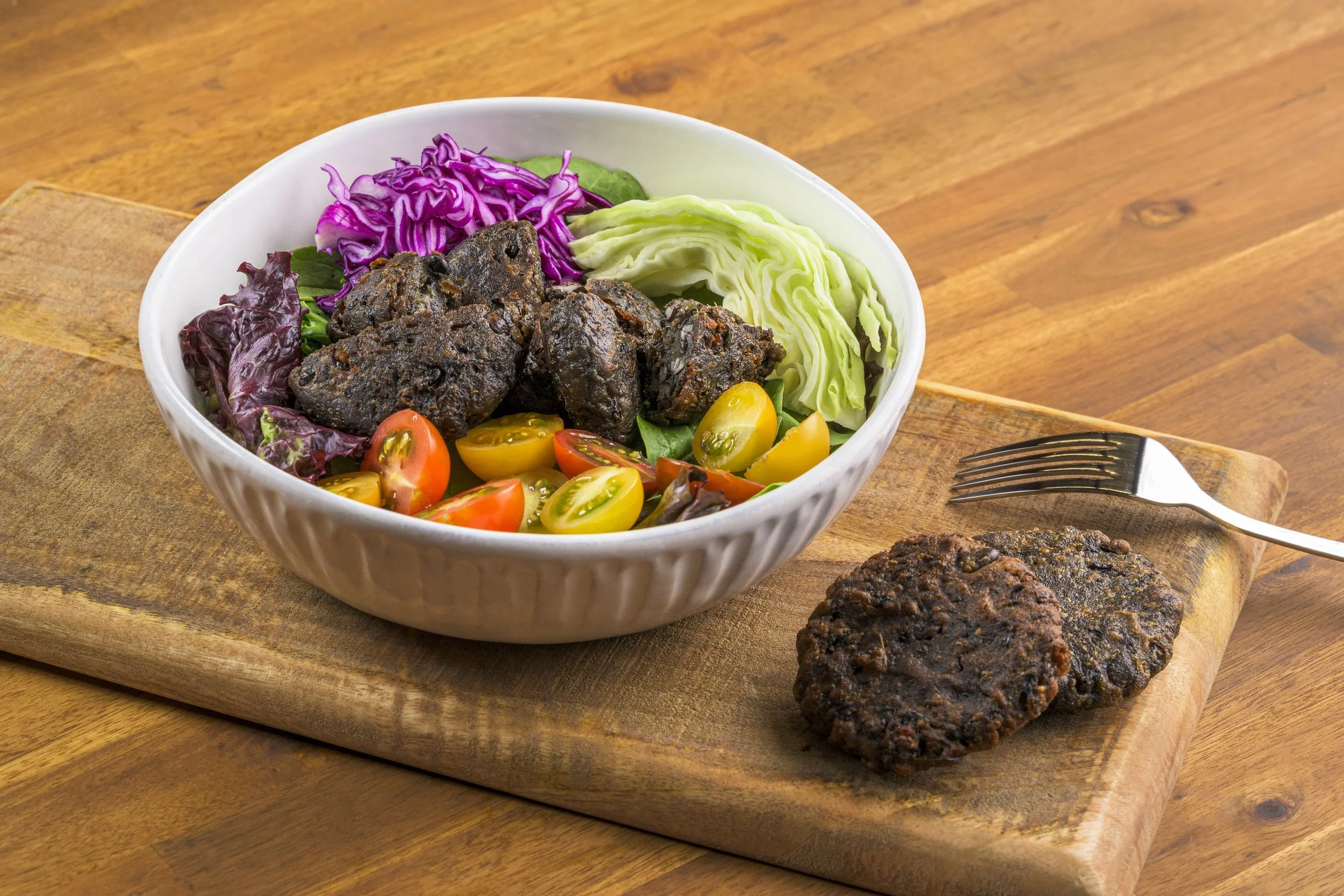 A white bowl of salad with purple cabbage, green lettuce, cherry tomatoes, and dark meat chunks, placed on a wooden cutting board with a fork and two additional meat pieces nearby.