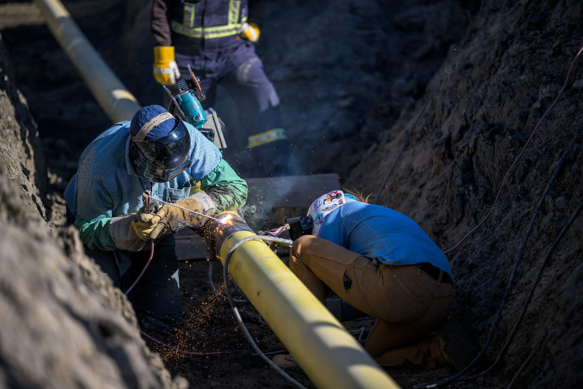 Workers welding a large yellow pipeline in an outdoor trench with sparks flying and rocks surrounding them.
