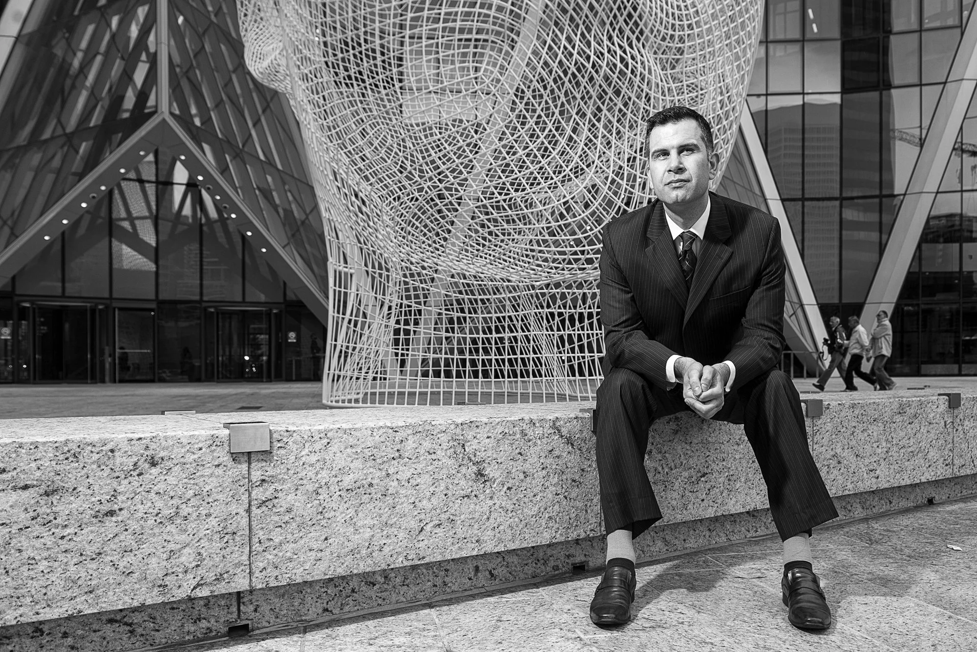 A man in a suit sitting on a concrete ledge outside a modern building with a large, intricate metal sculpture behind him.