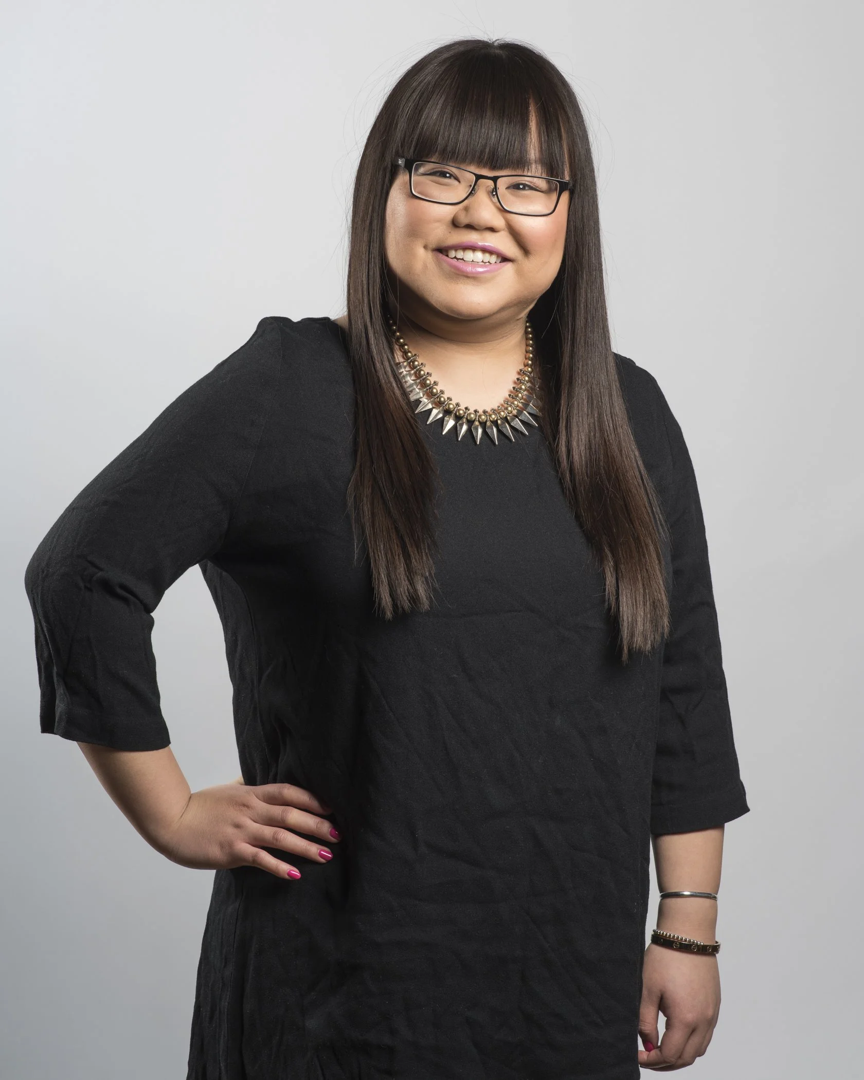 A woman with straight dark hair, wearing glasses, a black dress, and a spiked necklace, smiling and posing with her hand on her hip against a plain white background.