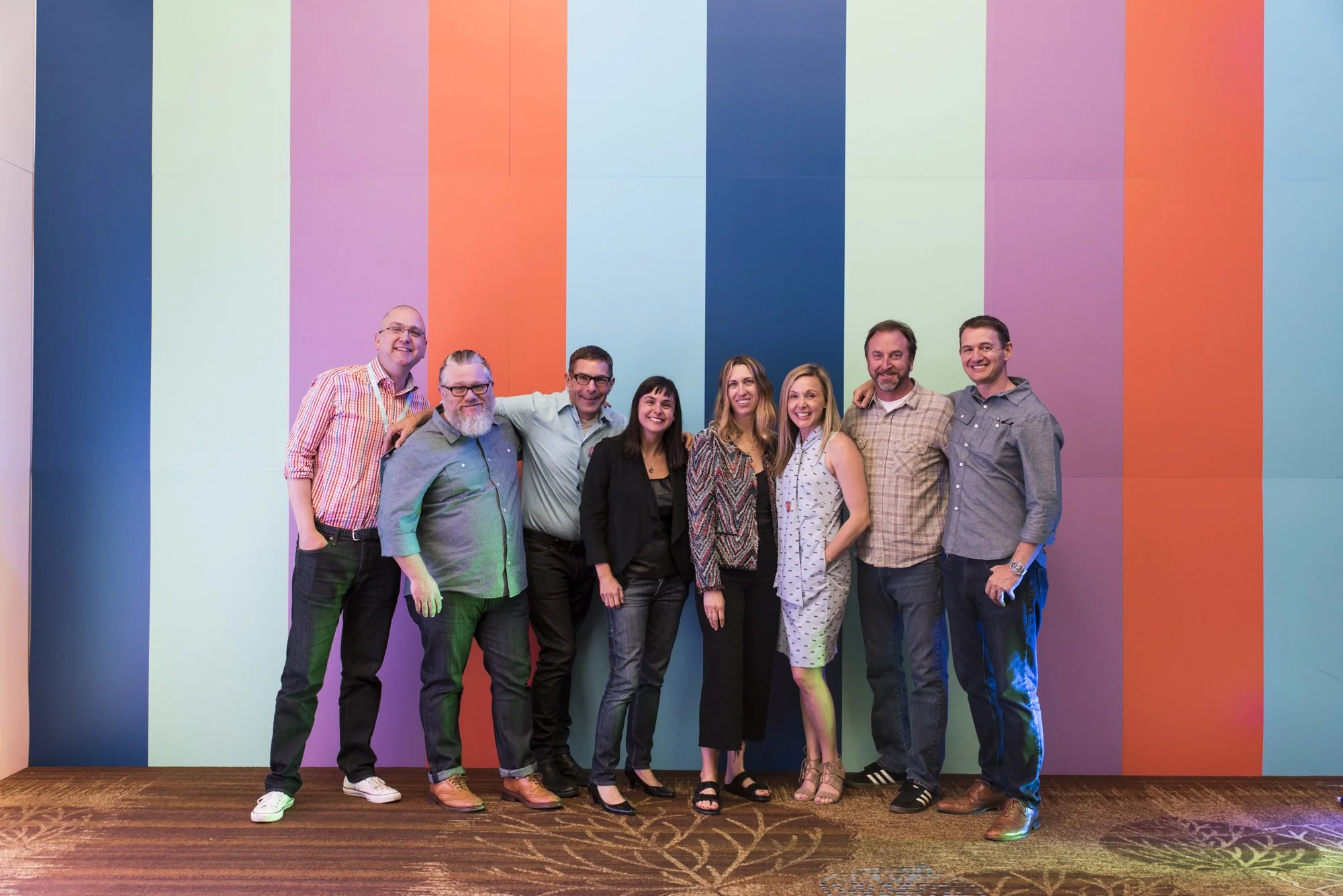 Group of nine people standing together in front of a colorful striped wall, smiling, with arms around each other.