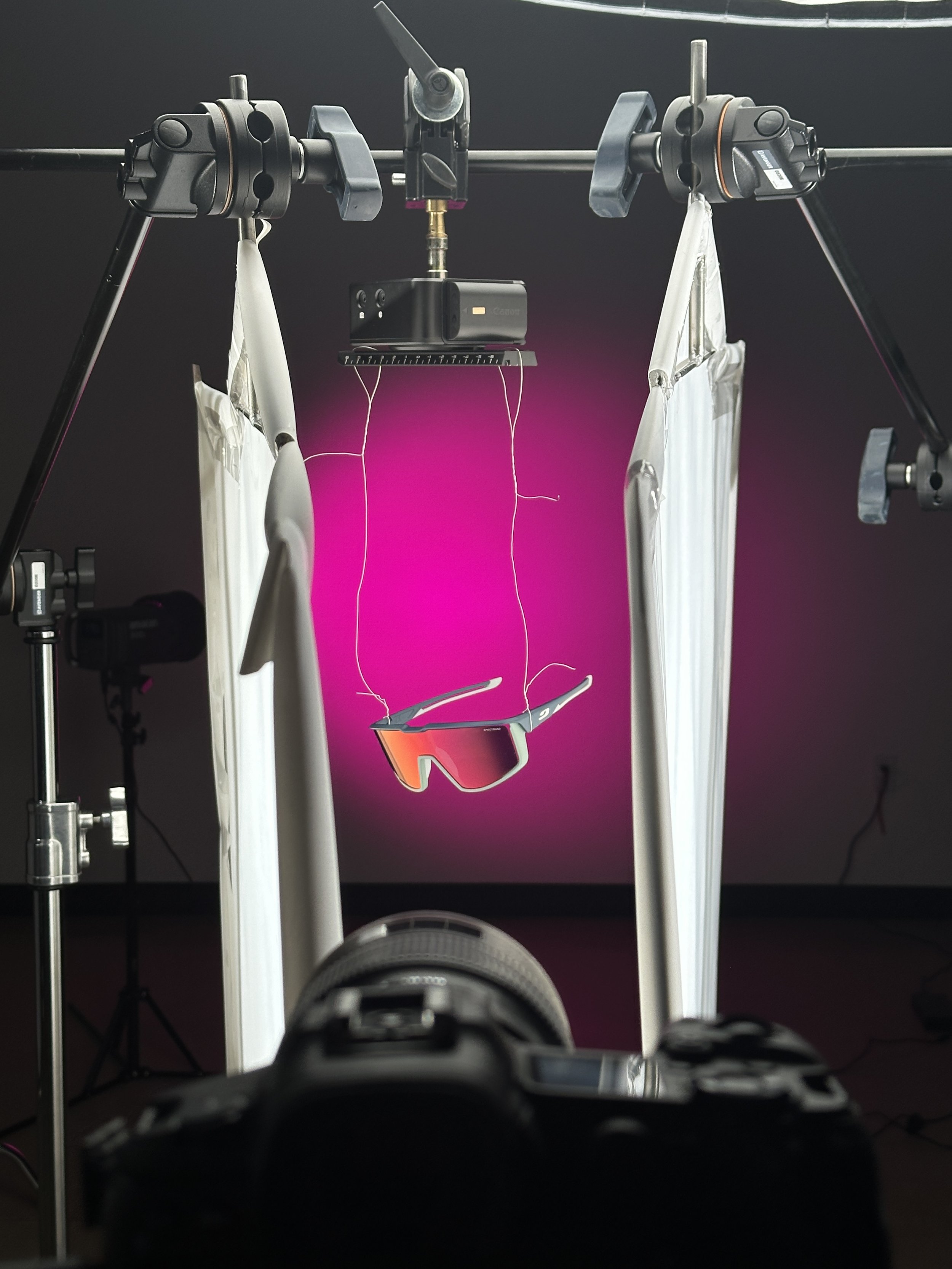 Set up for product photography featuring sunglasses suspended in front of a purple pink gradient background, with professional lighting, support stands, and a camera in the foreground.