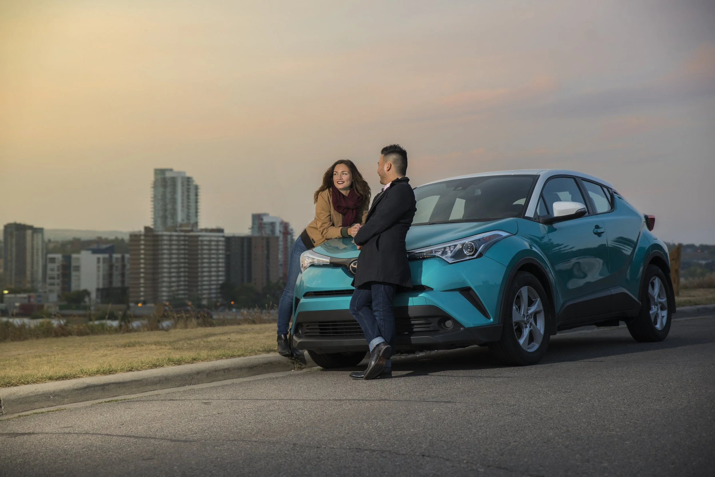 A man and a woman stand near a blue crossover car on a roadside, talking and smiling during sunset in an urban area with tall buildings in the background.