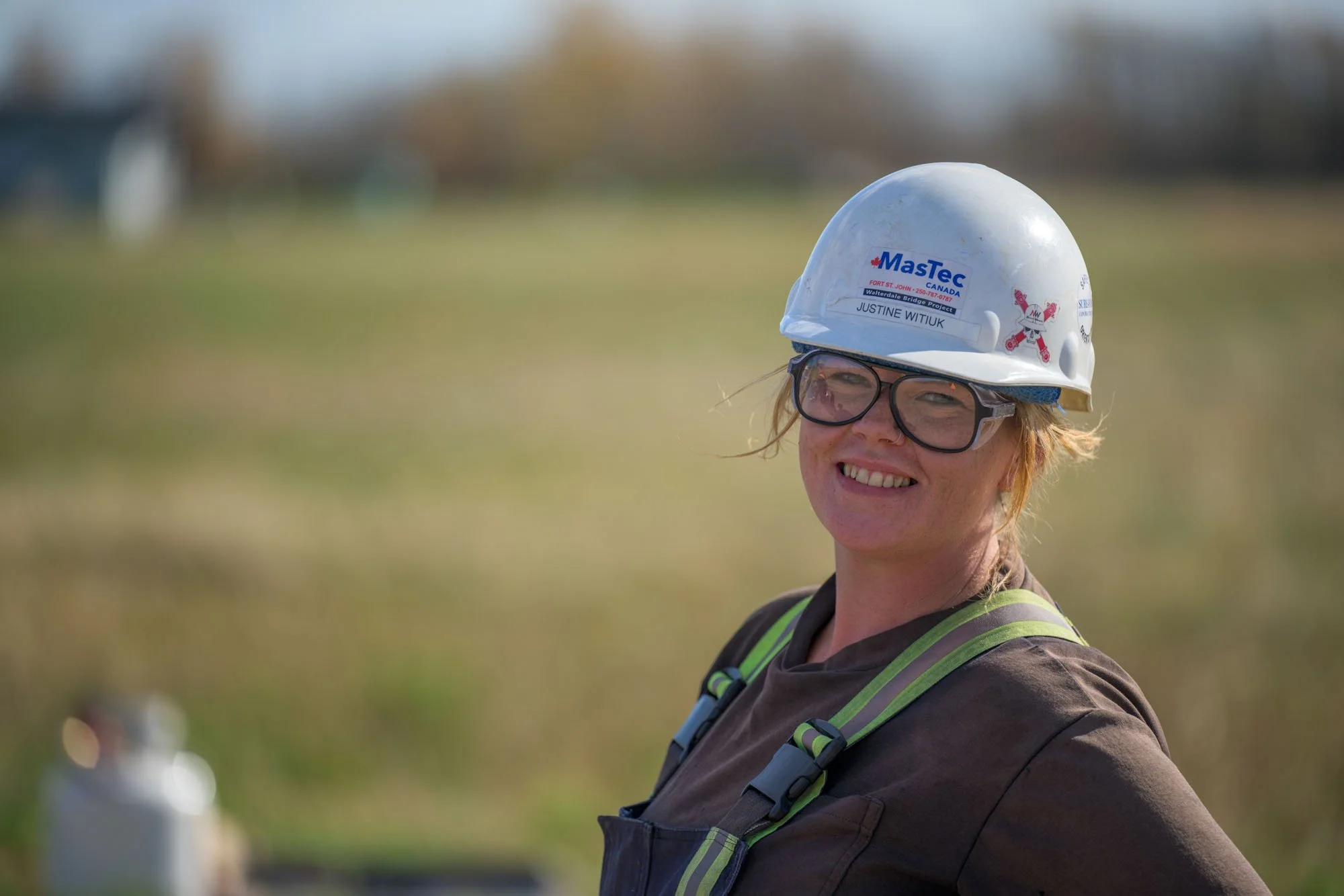 A smiling woman wearing safety glasses and a white safety helmet with logos, outdoors on a sunny day.