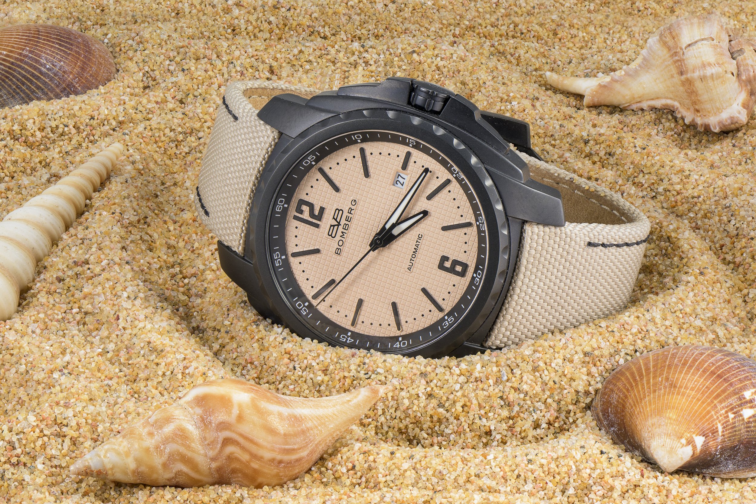 A beige-faced Bombard automatic wristwatch with black case, placed on sandy beach surrounded by seashells.