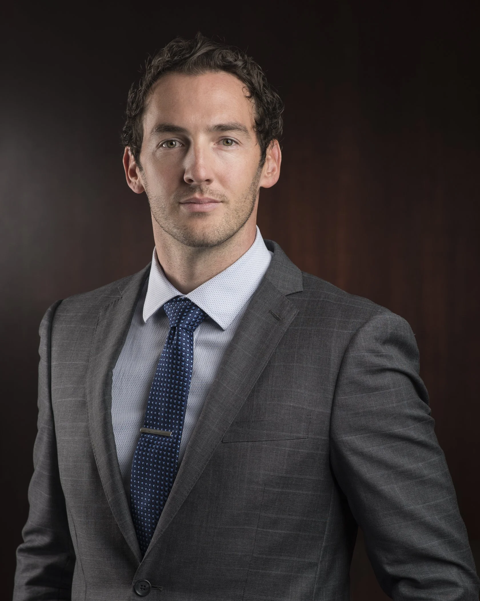 Professional portrait of a man wearing a gray business suit, light blue dress shirt, and navy blue patterned tie, standing against a dark gradient background.