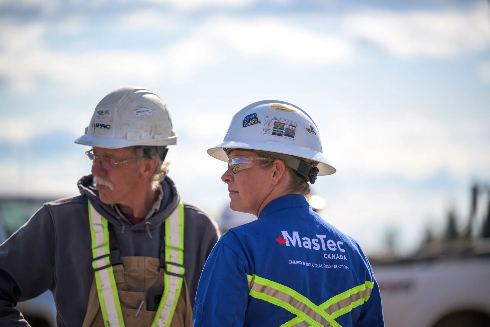 Two construction workers wearing safety helmets and reflective vests, one with a MaTec Canada logo on her jacket, standing outdoors during daytime with a cloudy sky in the background.