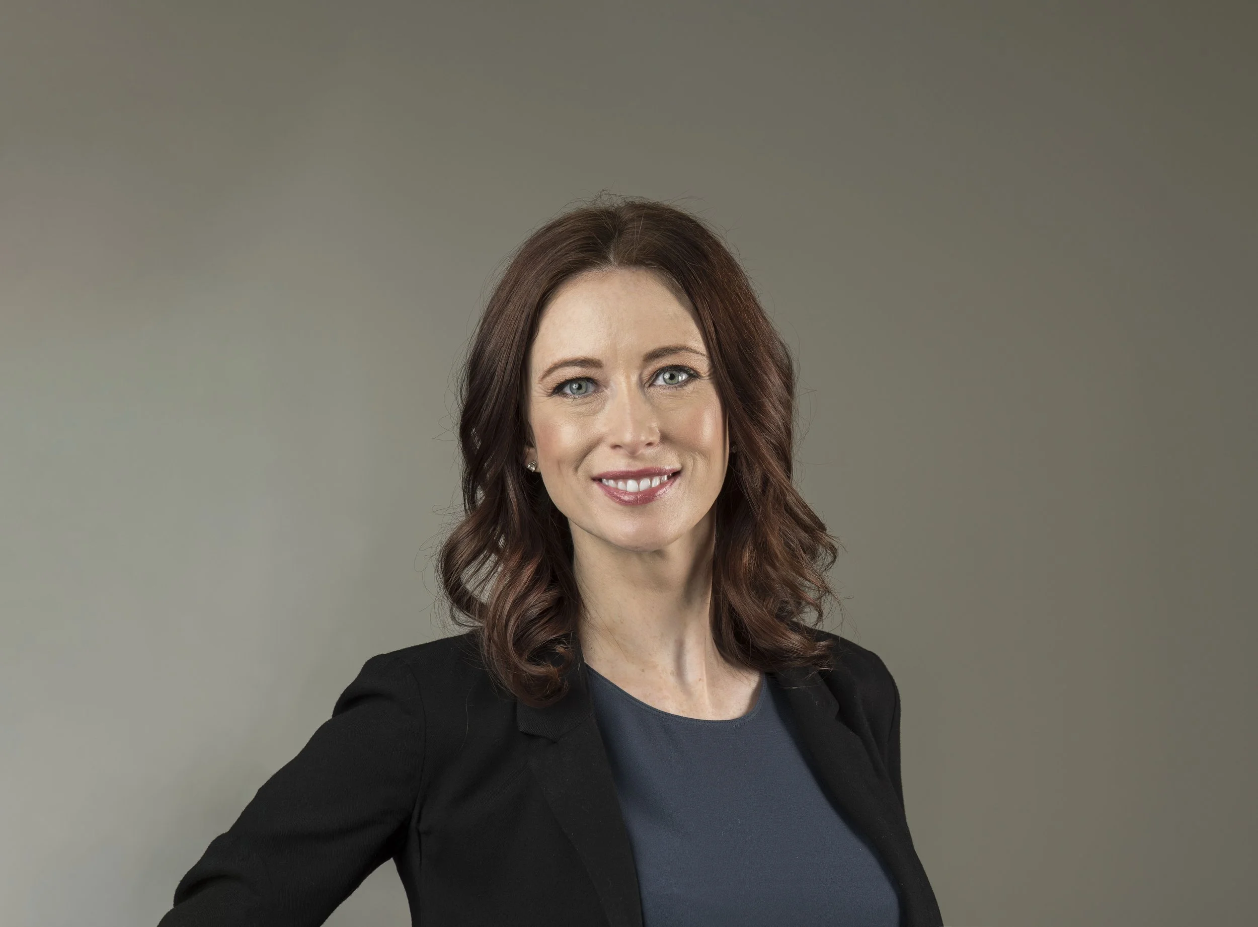 Portrait of a woman with shoulder-length brown hair, blue eyes, wearing a black blazer and a blue top, smiling against a neutral background.
