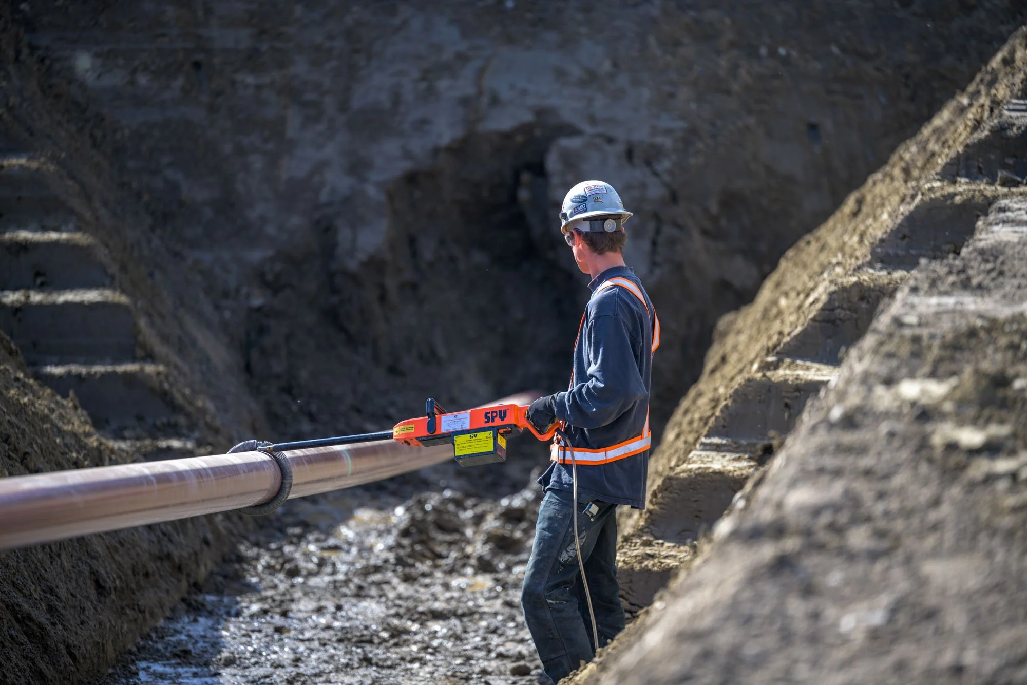 A construction worker in a hard hat and safety vest inspecting or working on a large underground pipeline in a trench.