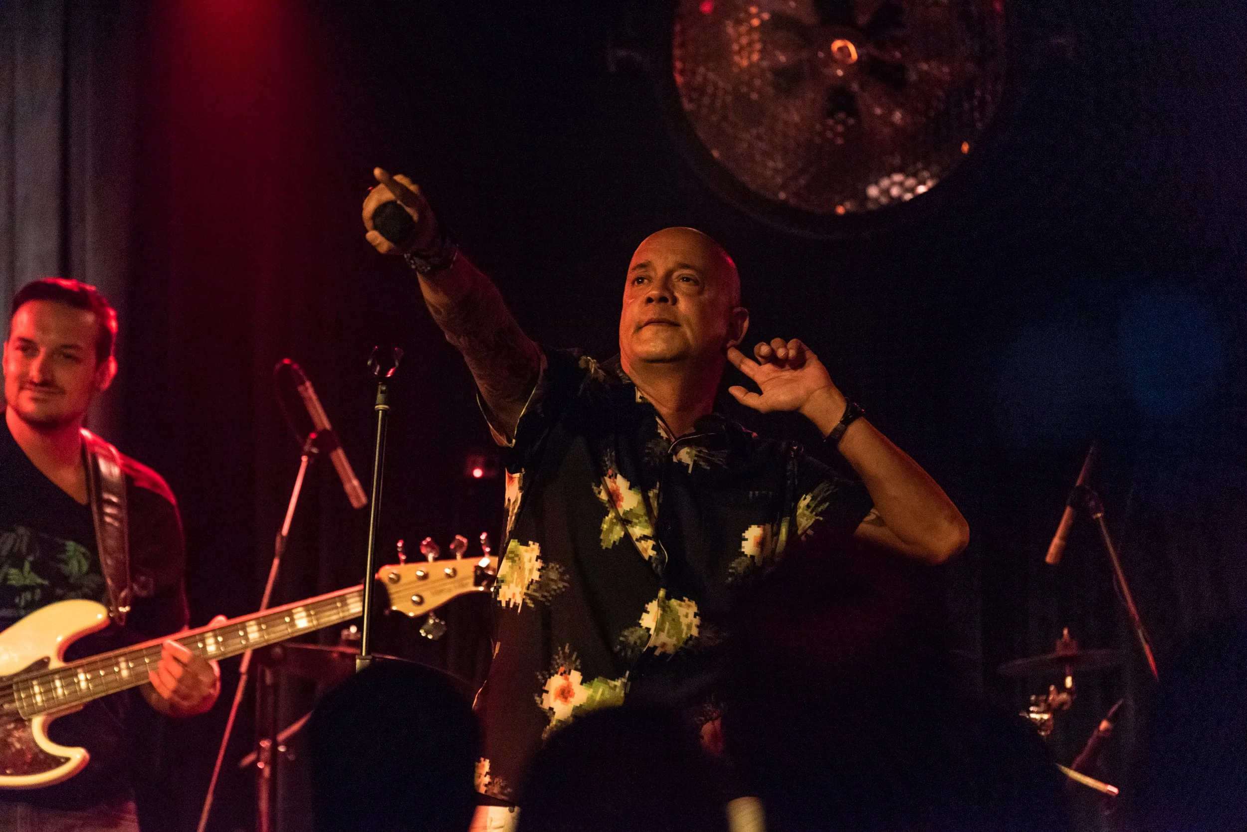 Man with a bald head wearing a colorful floral shirt is on stage, holding a microphone close to his head and pointing with his right hand. Another man with a guitar is on the left, smiling. The stage has black curtains and a large disco ball hanging 