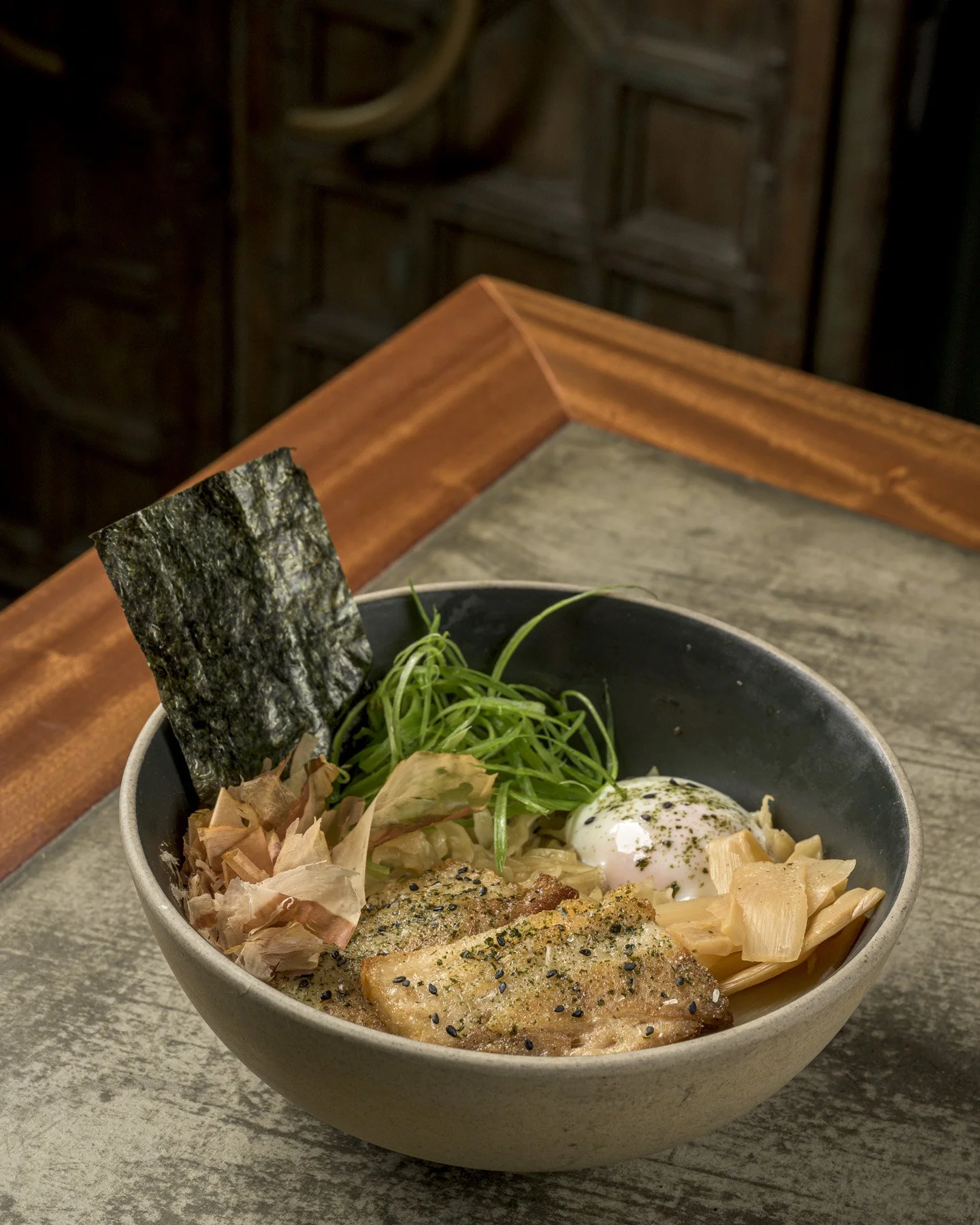A bowl of ramen with grilled fish, seaweed, green onions, soft boiled egg, bamboo shoots, and bonito flakes on a rustic wooden table.