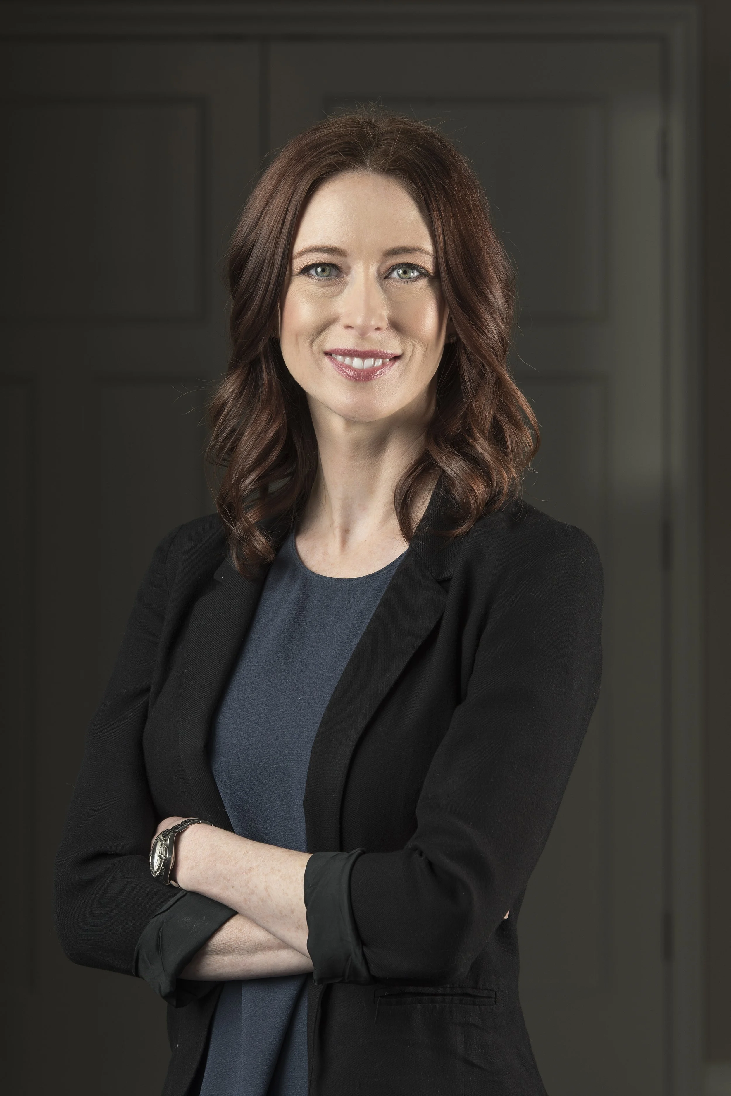 A woman with shoulder-length brown hair, wearing a dark blazer and blue top, smiling with crossed arms in front of a neutral background.