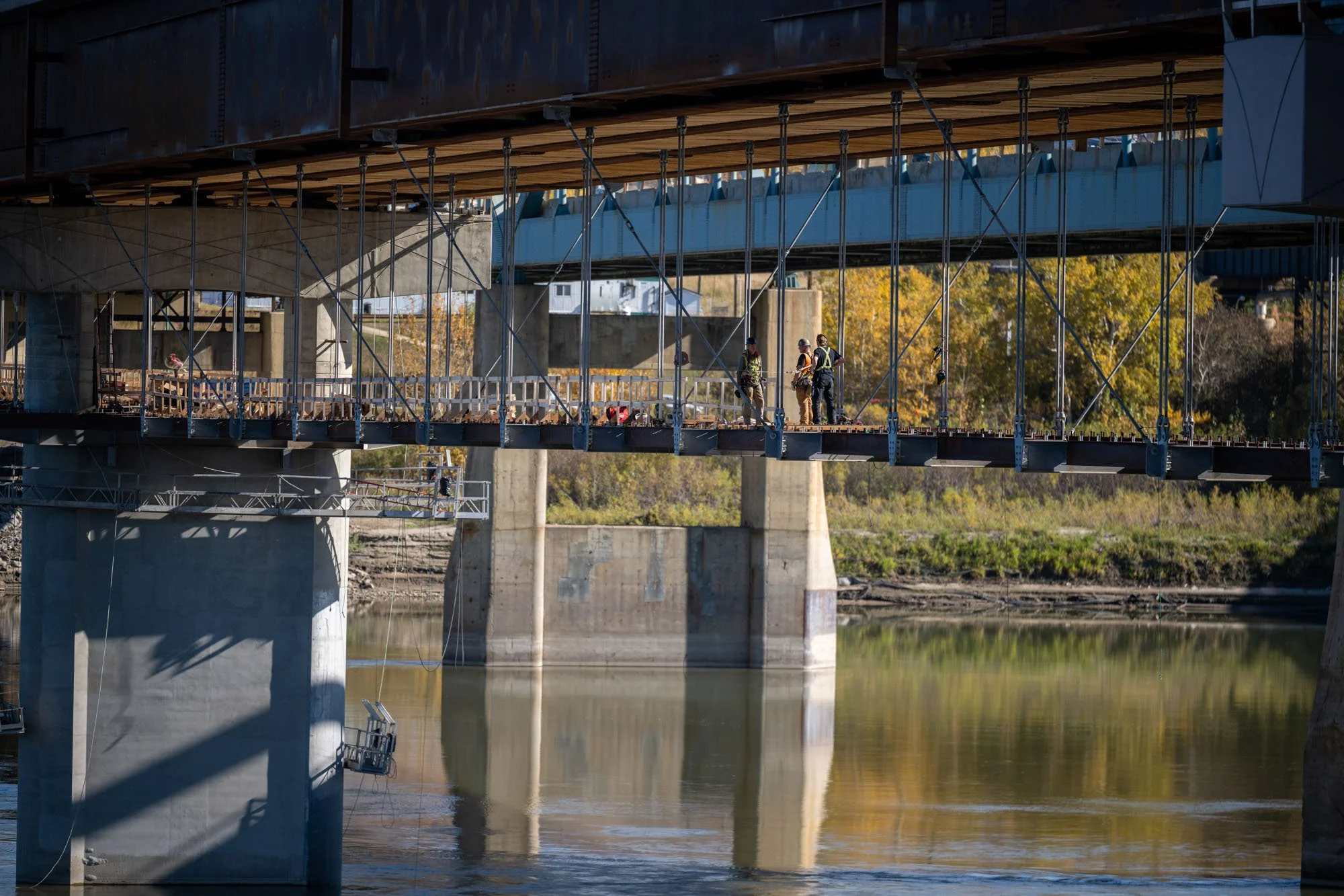 Construction workers standing on a suspended bridge under a large bridge over a river.