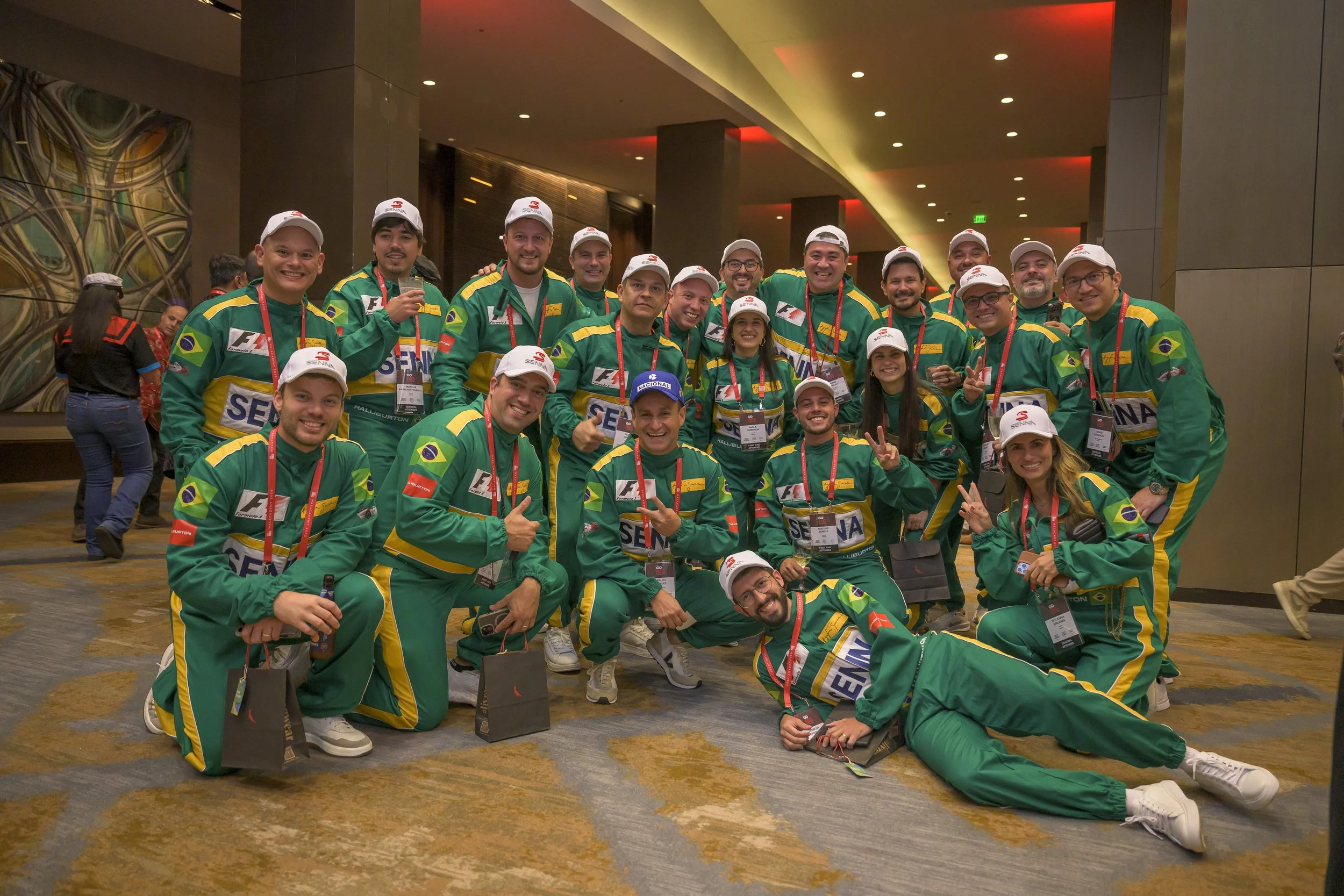A group of people in matching green, yellow, and blue racing suits with Brazil flags, posing for a photo at an indoor event. Many are making peace signs or thumbs-up gestures, and some wear white caps with red logos.