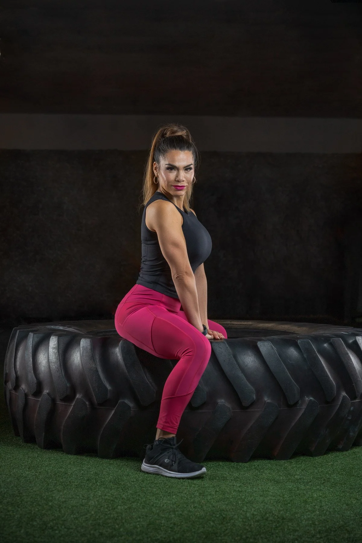A woman sitting on a large tractor tire in a gym, wearing a sleeveless black top, bright pink leggings, and black athletic shoes, with her hair in a high ponytail and a focused expression.