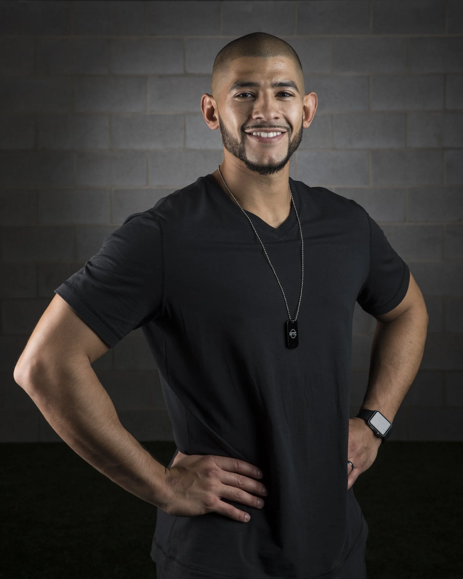 A smiling man with a beard and short hair, wearing a black athletic shirt, a necklace with a dog tag, a smartwatch, and a ring, standing with hands on hips against a dark background.
