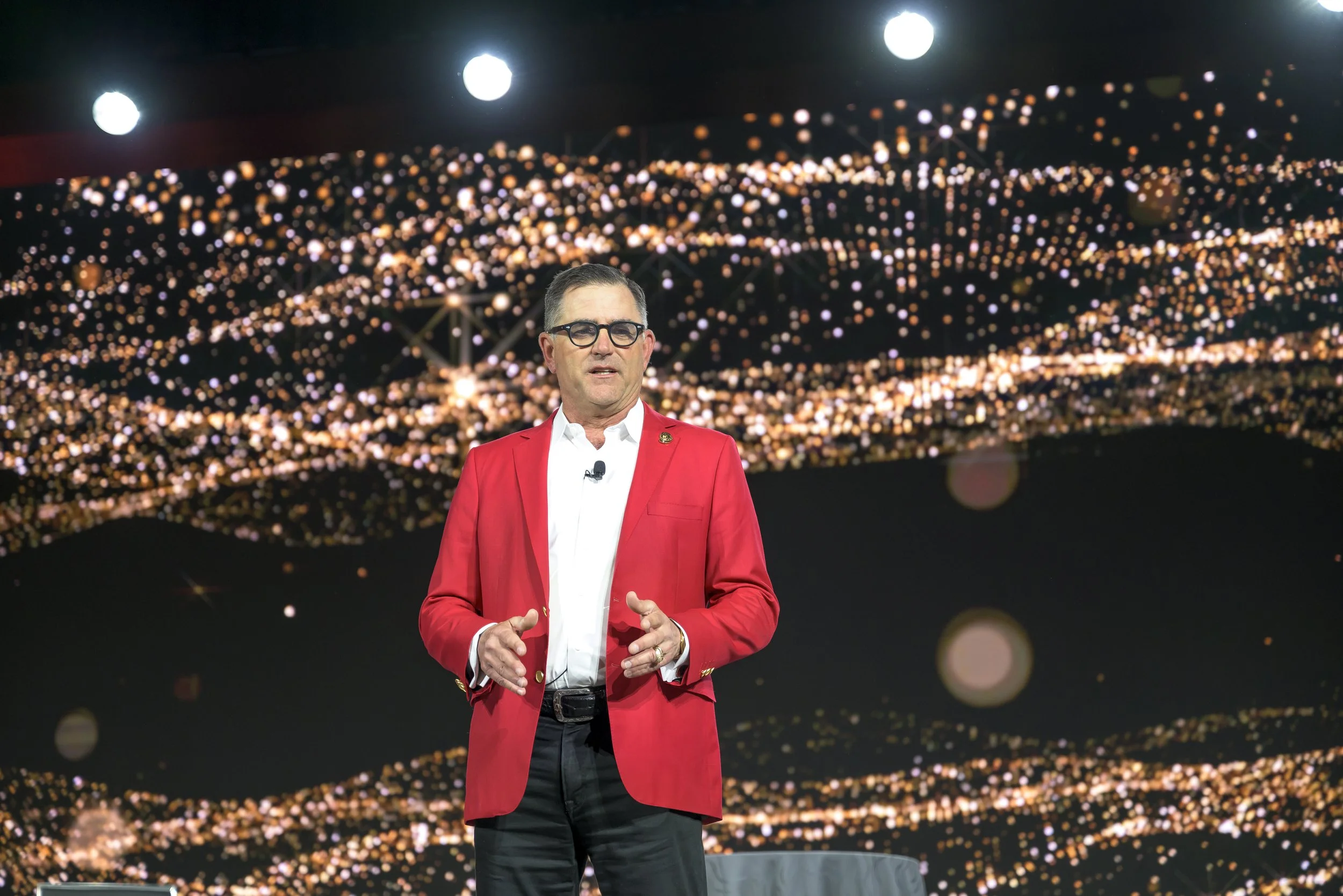 Man in red blazer speaking on stage with a sparkling, gold and black background.
