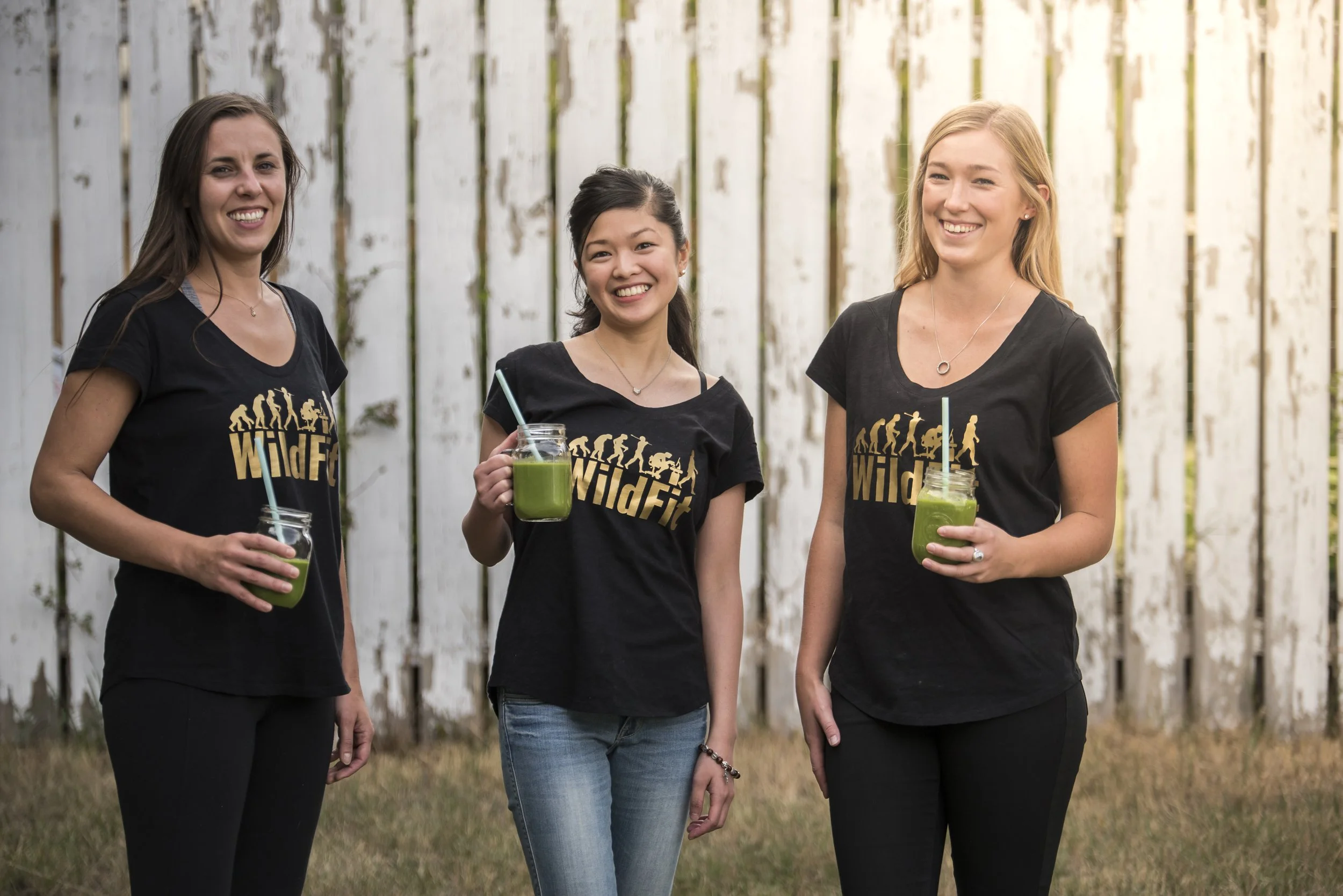 Three women smiling, standing outdoors in front of a white wooden fence, each holding a green beverage in a mason jar with a straw, wearing matching black T-shirts with a yellow logo that says 'WildFit'.