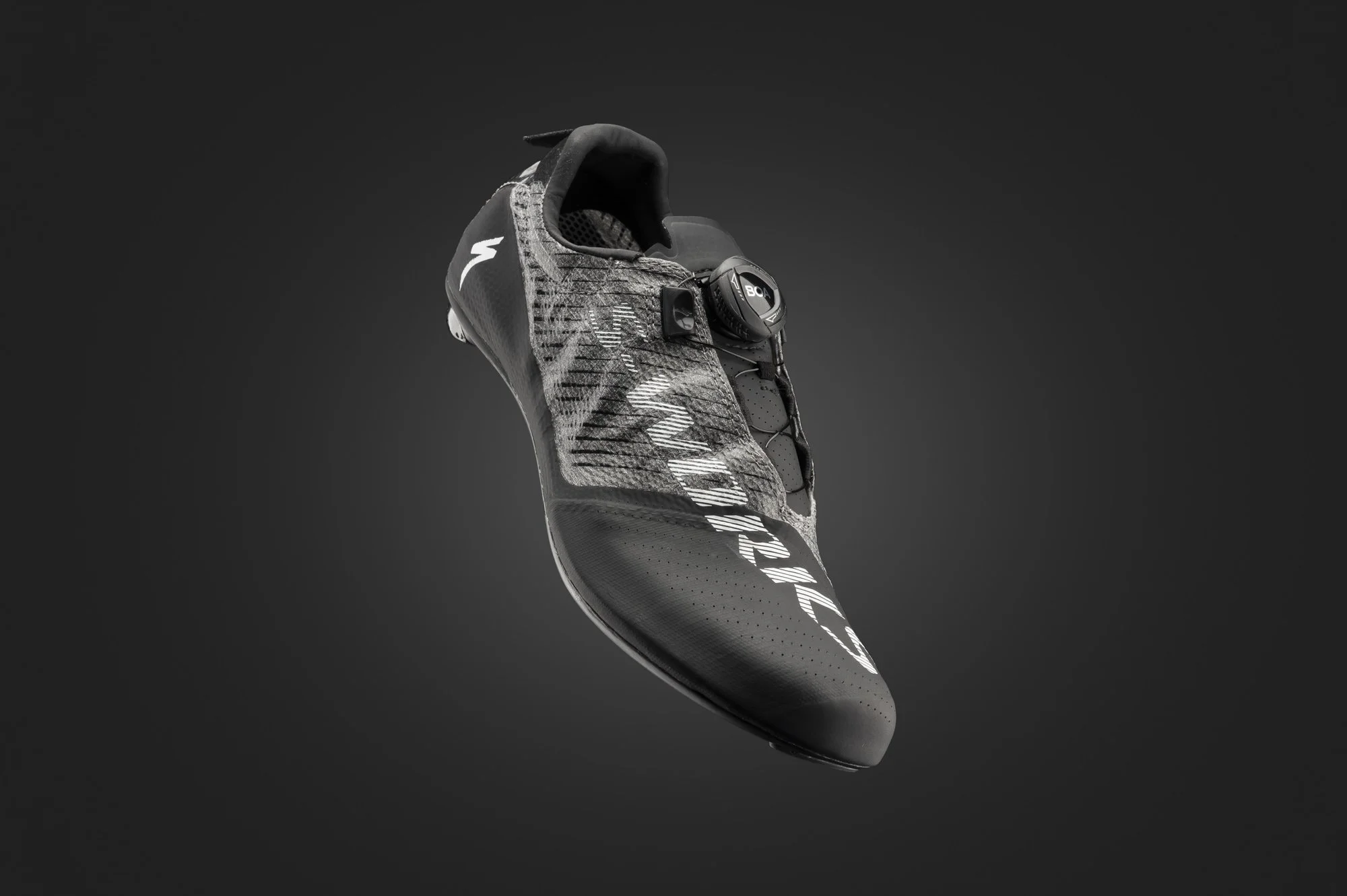 Close-up of a black and gray cycling shoe with a digital dial closure system, suspended against a dark background.